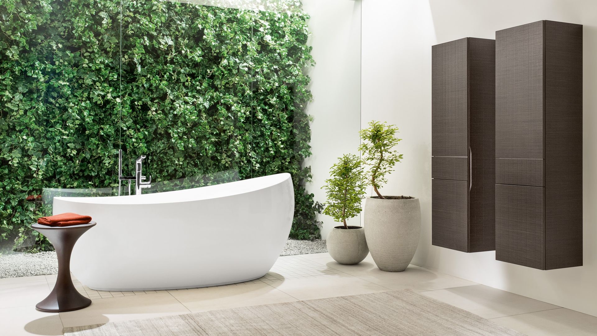 Baño con bañera exenta, ventanal con salida al jardín, muebles suspendidos de madera oscura, alfombra, taburete y macetas con plantas