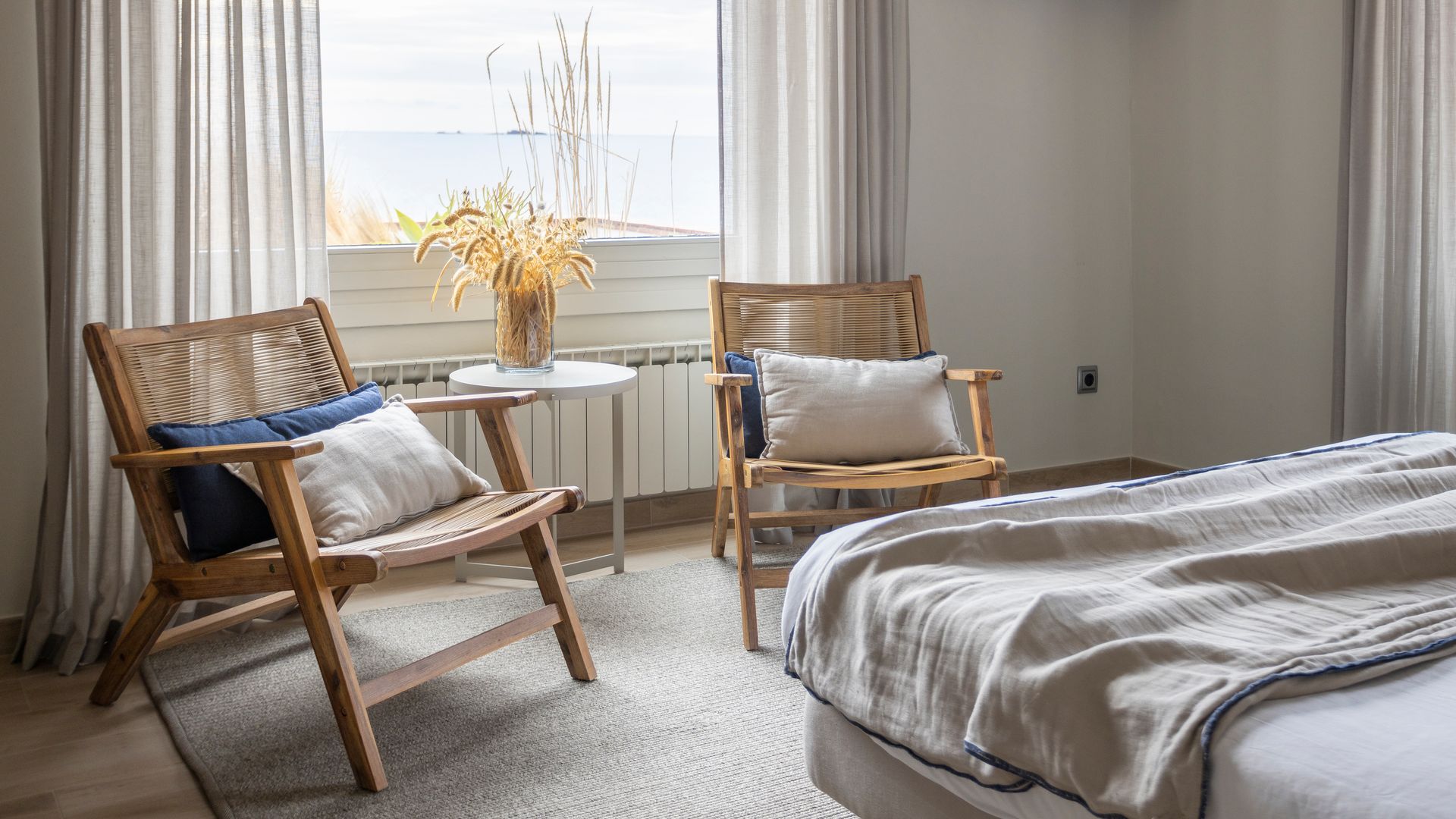 Dormitorio con gran ventanal, alfombra y ropa de cama de lino, rincón de lectura con con butacas y mesa auxiliar, cortinas beiges