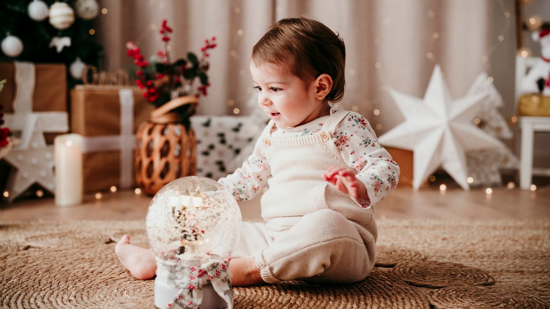 Bebé jugando con una bola de Navidad en el suelo de una casa decorada llena de regalos.