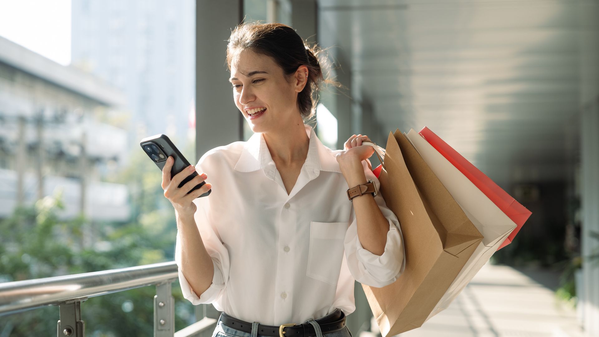 Mujer joven con bolsas de la compra utilizando el teléfono móvil por la calle de una ciudad, con ropa de primavera-verano.