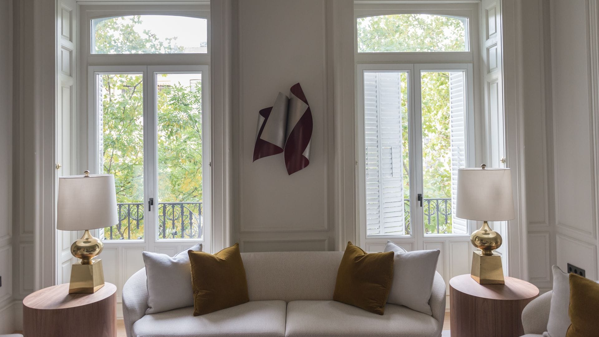 Salón con sofá blanco, ventanas con contraventanas de madera blanca, cojines, lámparas de sobremesa con el pie dorado 