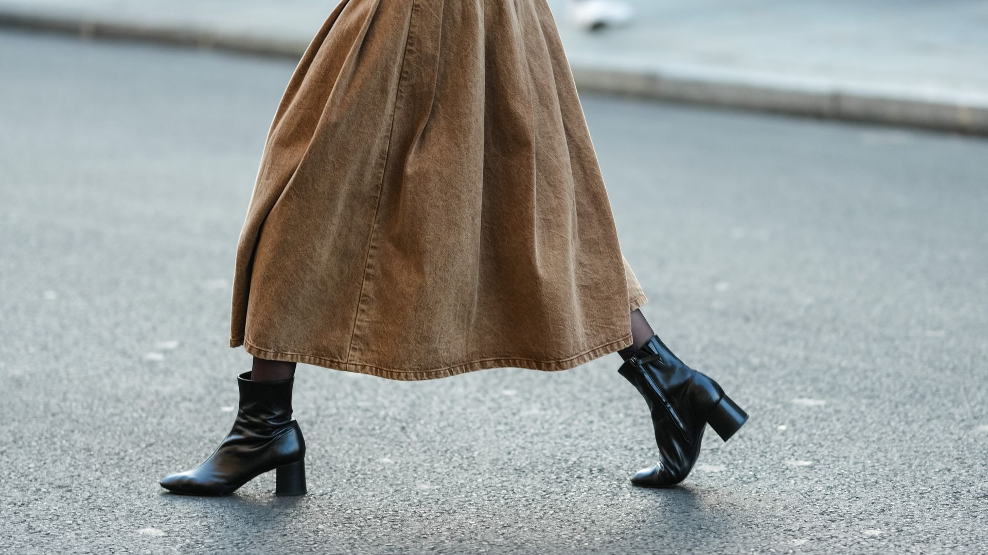 Mujer caminando por el asfalto cruzando una calle con botines de piel negra y abrigo largo en marrón. 