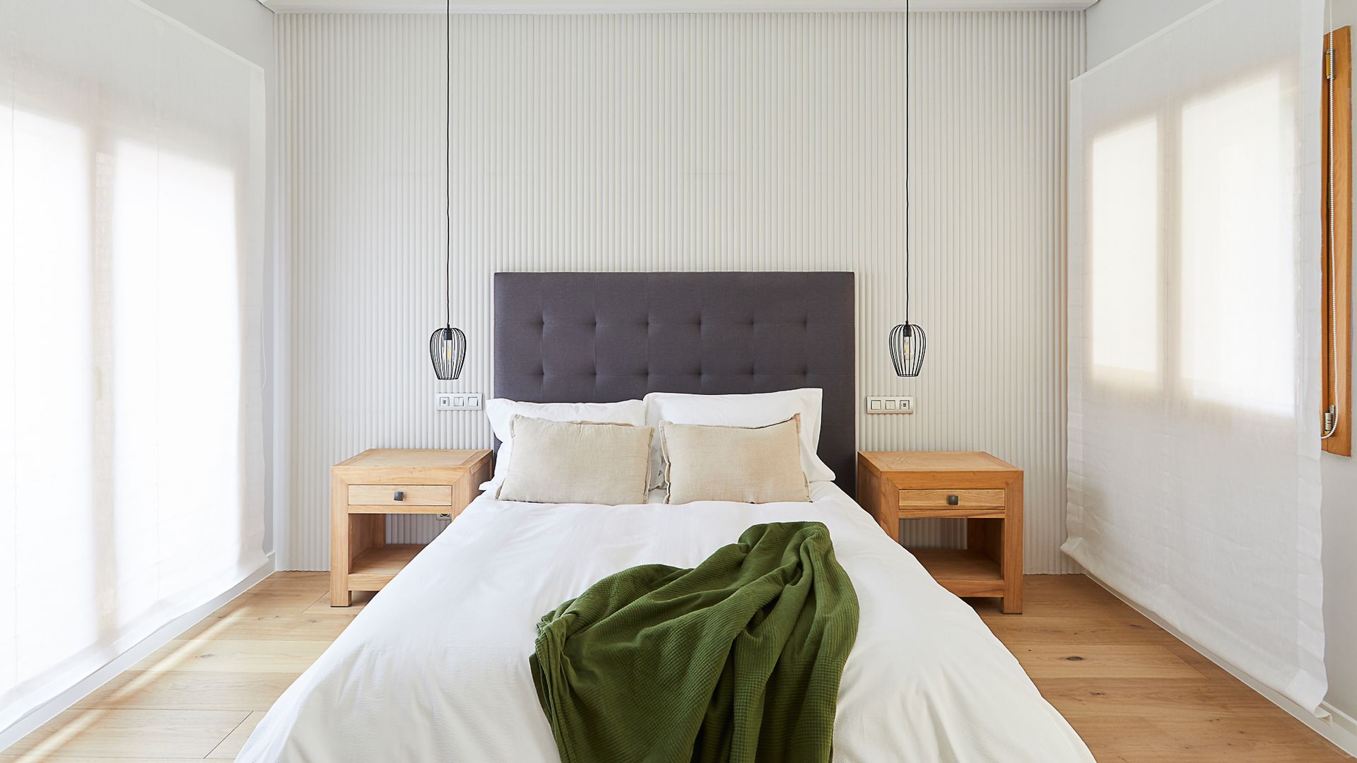 Dormitorio con pared del cabecero con papel pintado, cabecero tapizado en capitoné gris oscuro, estores blancos hasta el suelo, mesitas de noche de madera, lámparas de techo a ambos lados de la cama 