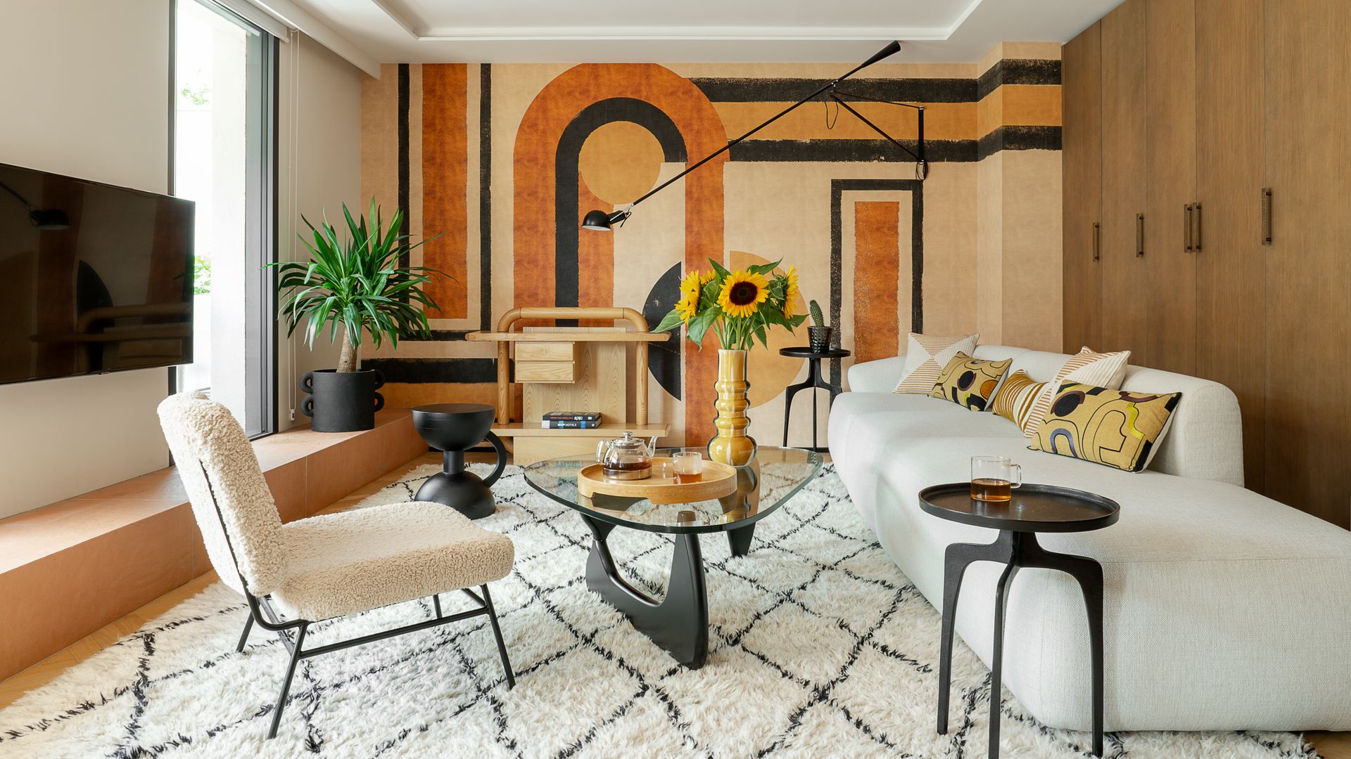Salón con mural de papel pintado, sofá blanco con respaldo bajo, alfombra de pelo, mesa de centro Coffee de Isamu Noguchi, taburete negro, butaca, jarrón con girasoles
