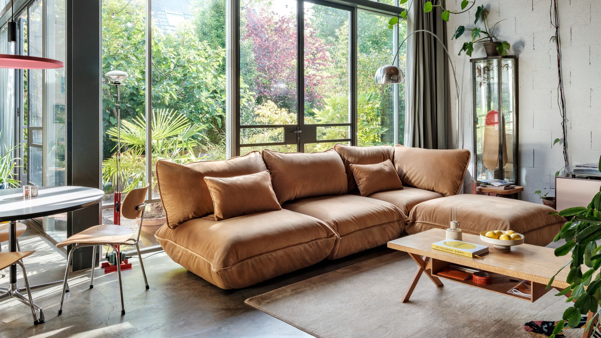 Salón con grandes ventanales con salida al jardín, sofá con chaise longue de color marrón, mesa de centro de madera con bandeja inferior. alfombra, plantas colgantes, lámpara de pie tipo Arco