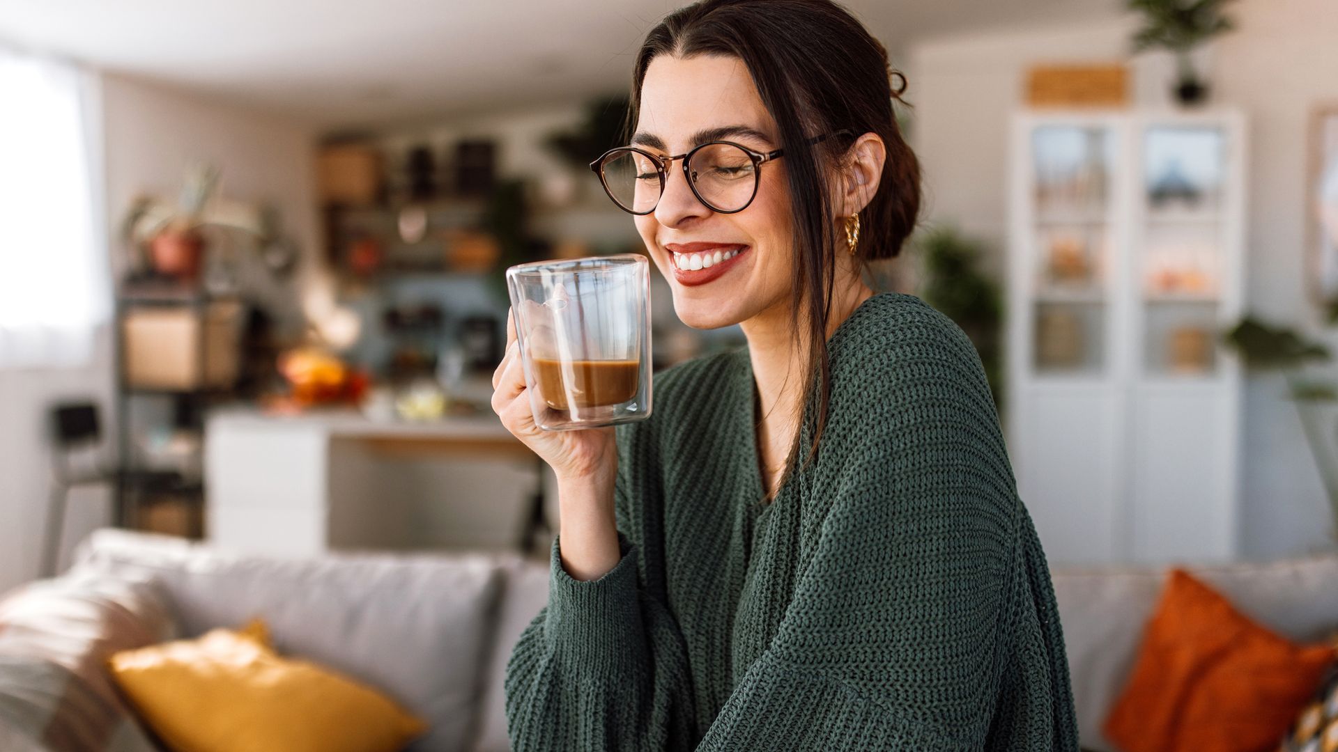 Una mujer sonriendo con ropa de otoño con un café en las manos, sentada en el sofá del salón de su casa, con la cocina por detrás.