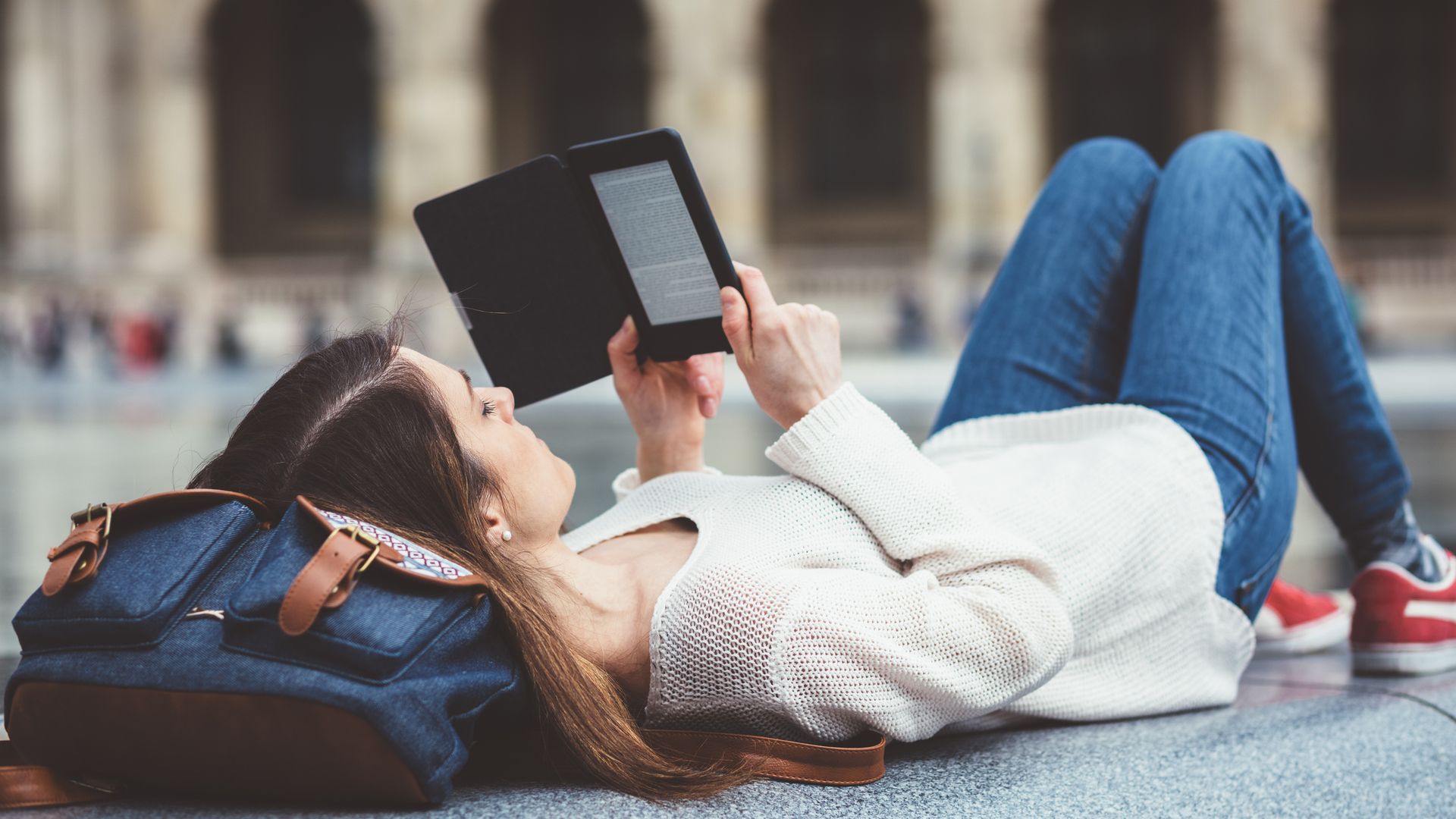 Chica joven con ropa de otoño-invierno tumbada en un banco de piedra de París leyendo en un libro electrónico con la cabeza apoyada en una mochila.