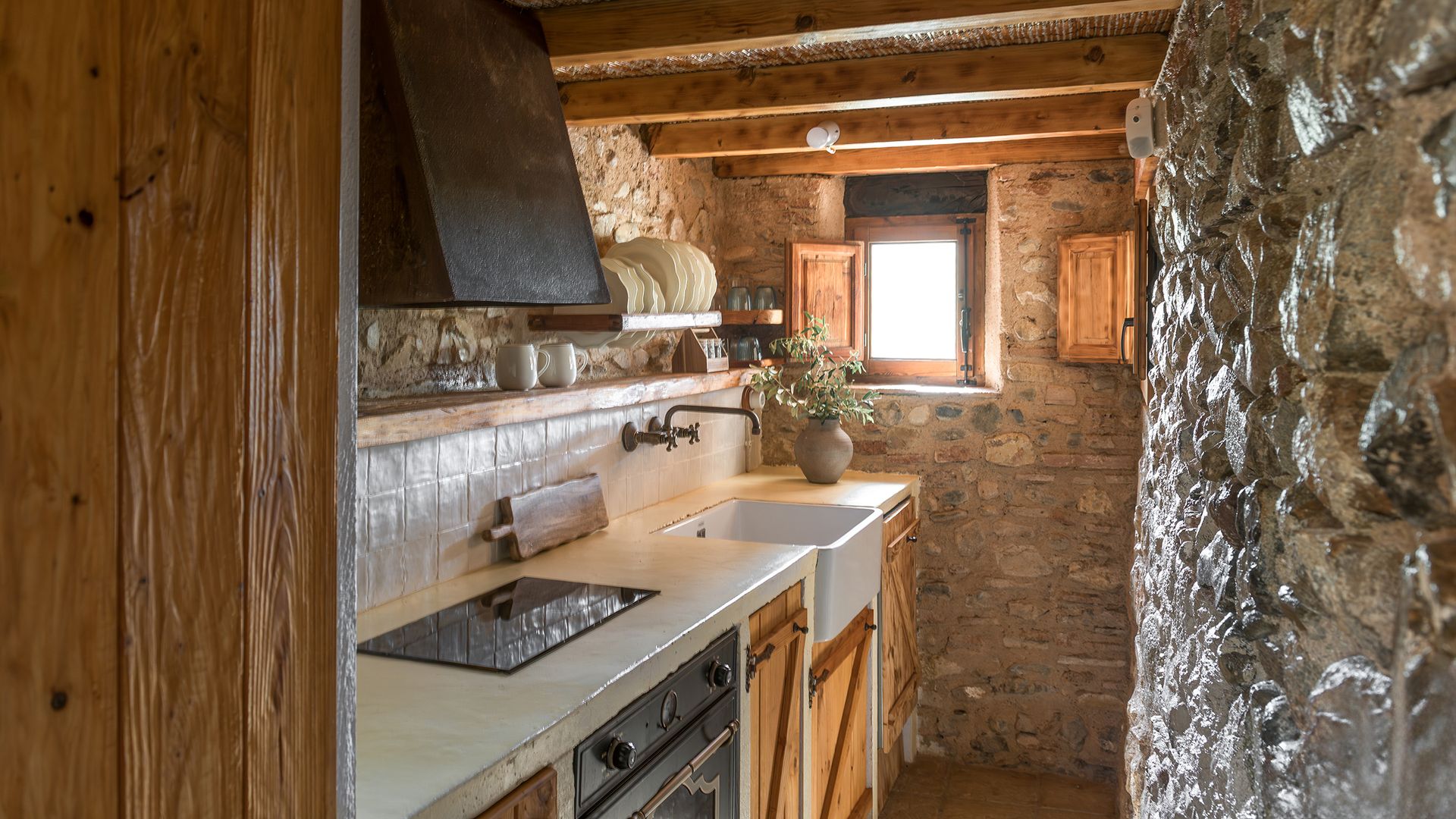 Cocina rústica con paredes de piedra, azulejos en la pared del antepecho, vigas de madera, balda de madera en lugar de muebles altos