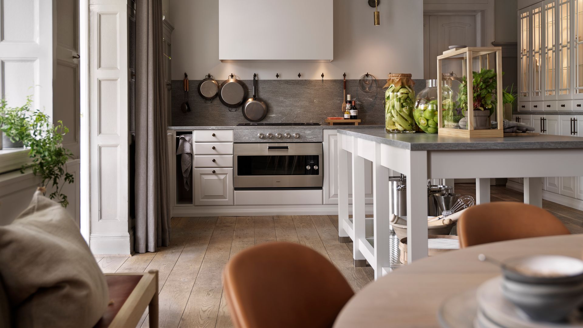 Cocina con mesa de trabajo blanca con sobre de piedra natural, sartenes colgadas en ganchos, tarros de cristal con alimentos, vivero de plantas 