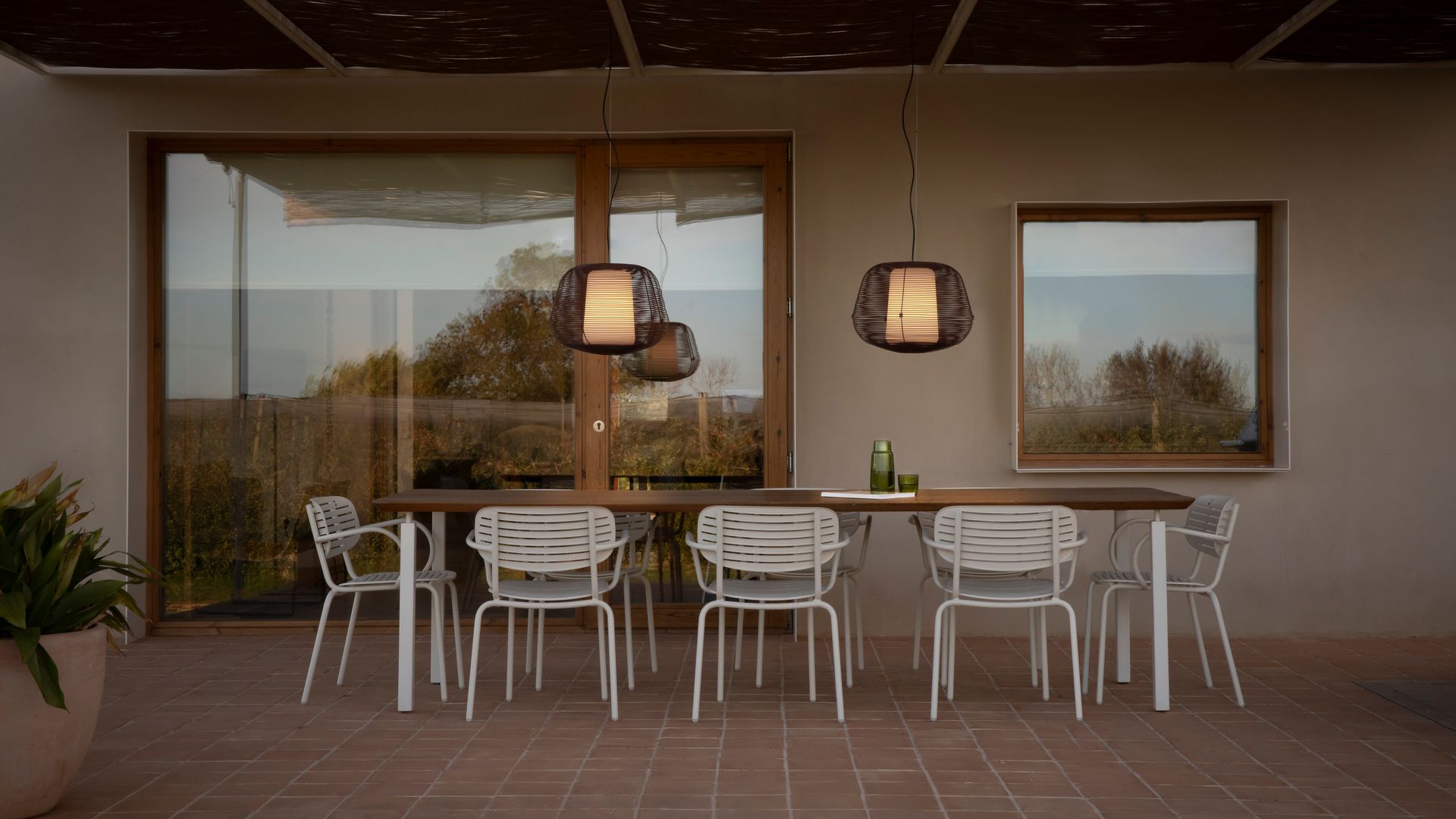 Porche con comedor con mesa de madera, sillas blancas, suelo de barro y lámparas de techo