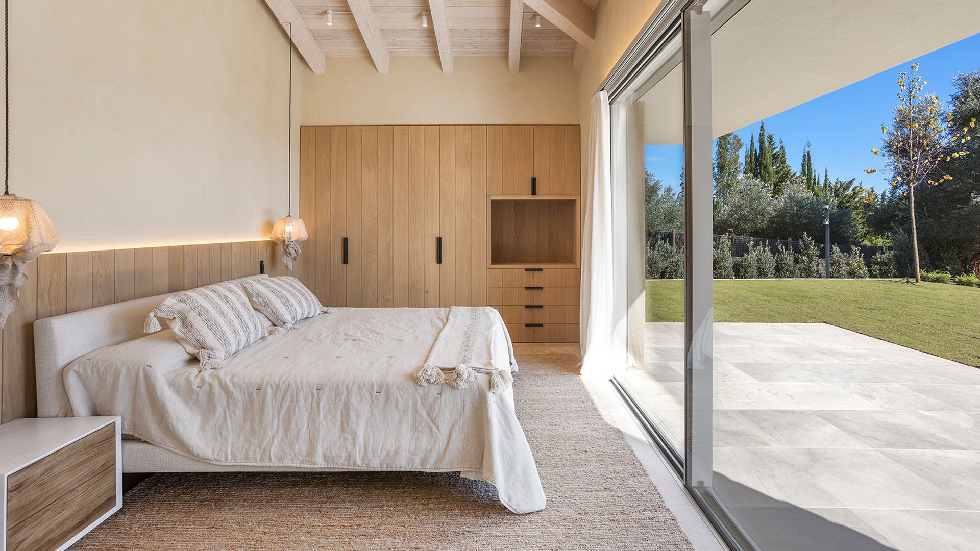 Dormitorio con salida al jardín, con friso de madera, cabecero tapizado, vigas en el techo, ropa de cama blanca, armario de madera 