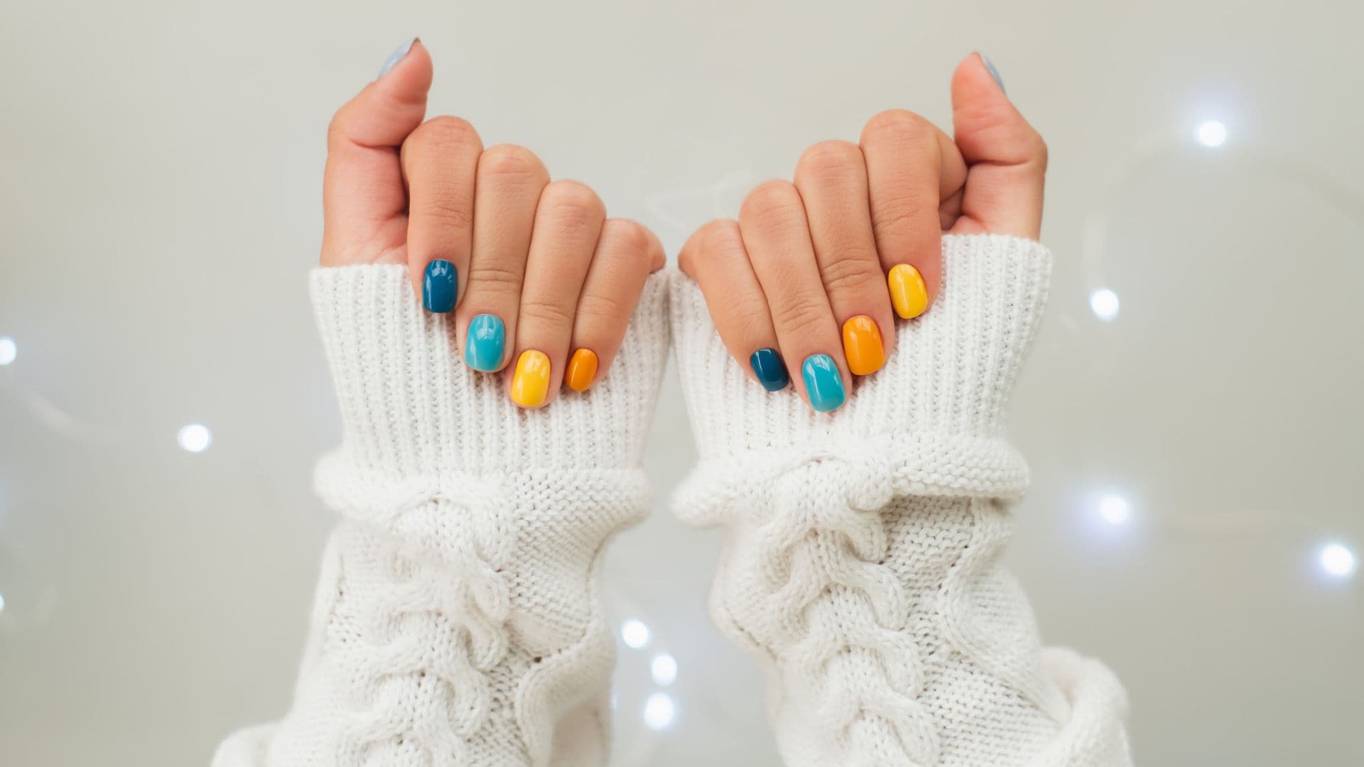 Manicura de colores en las manos de una mujer con jersey blanco y luces de Navidad al fondo en blanco.