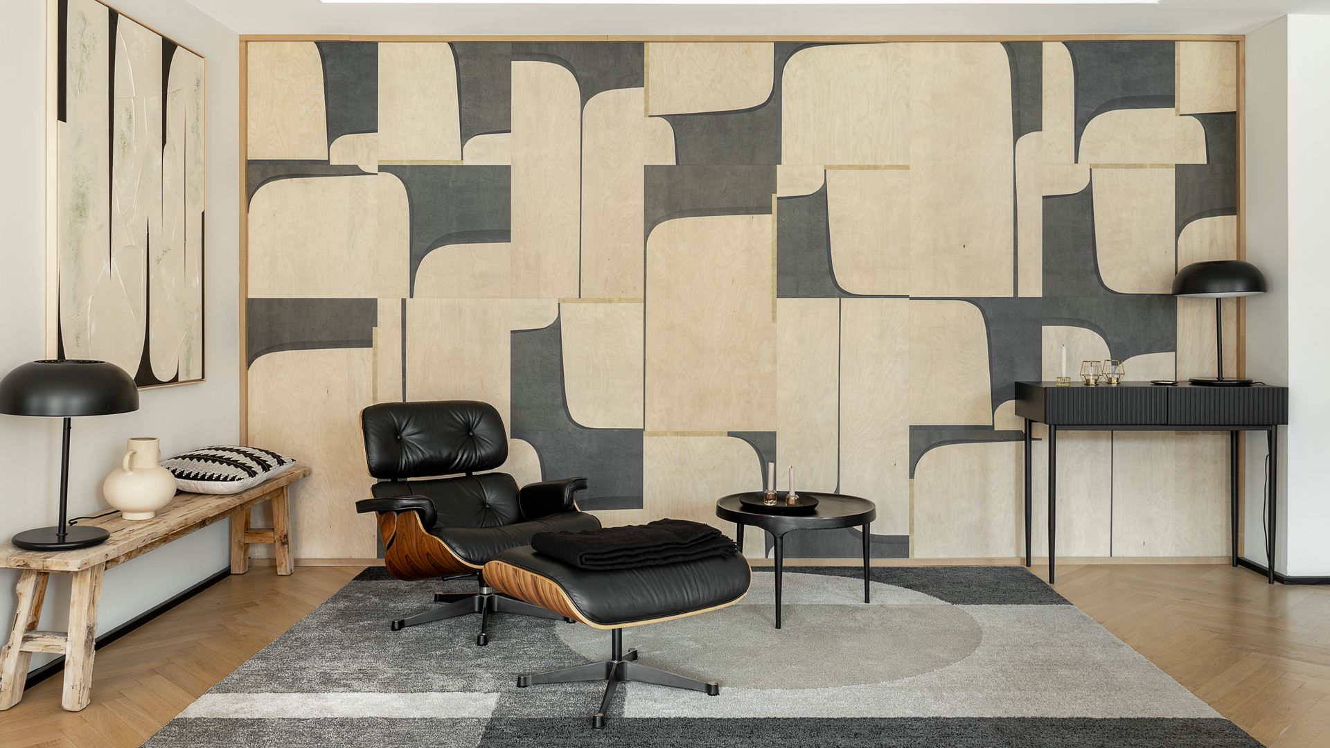 Rincón de lectura con Loungue Chair de los Eames, alfombra gris con motivos geométricos, pared con paneles en gris y beige, banco, consola, mesa auxiliar negra, lámpara de sobremesa negro, molduras en el techo
