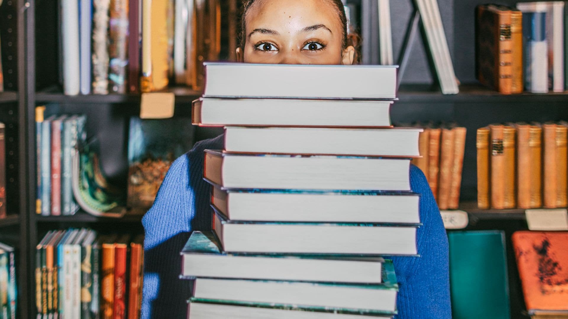 Chica joven sujetando una torre de libros entre sus manos, mirando a la cámara