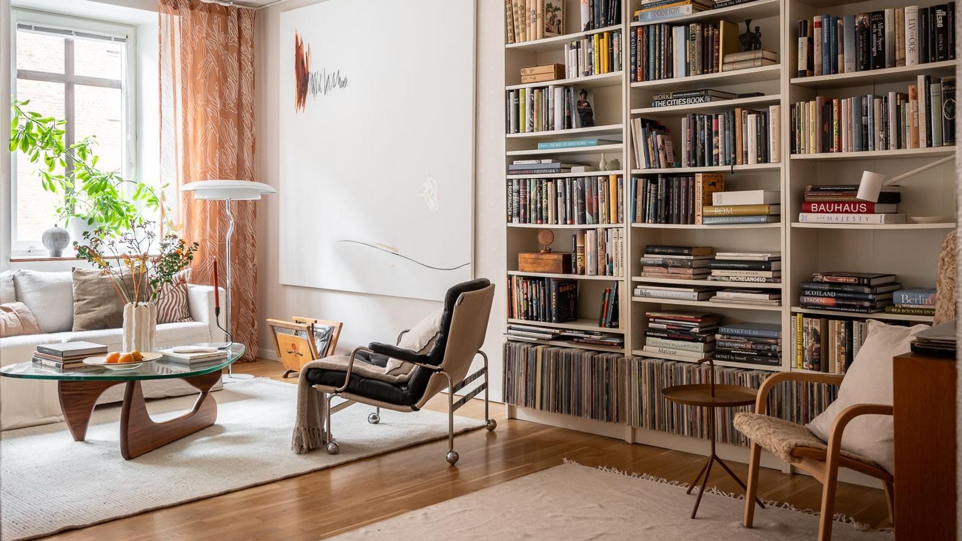 Salón con librería blanca llena de libros, rincón de lectura con butaca, mesa auxiliar, estar con mesa de centro con el sobre de cristal, lámpara de techo, alfombra, cuadro en la pared