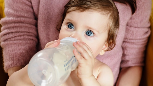 Una bebé de ojos azules bebiendo su leche en un biberón, mirando a la cámara en primer plano, en brazos de su madre