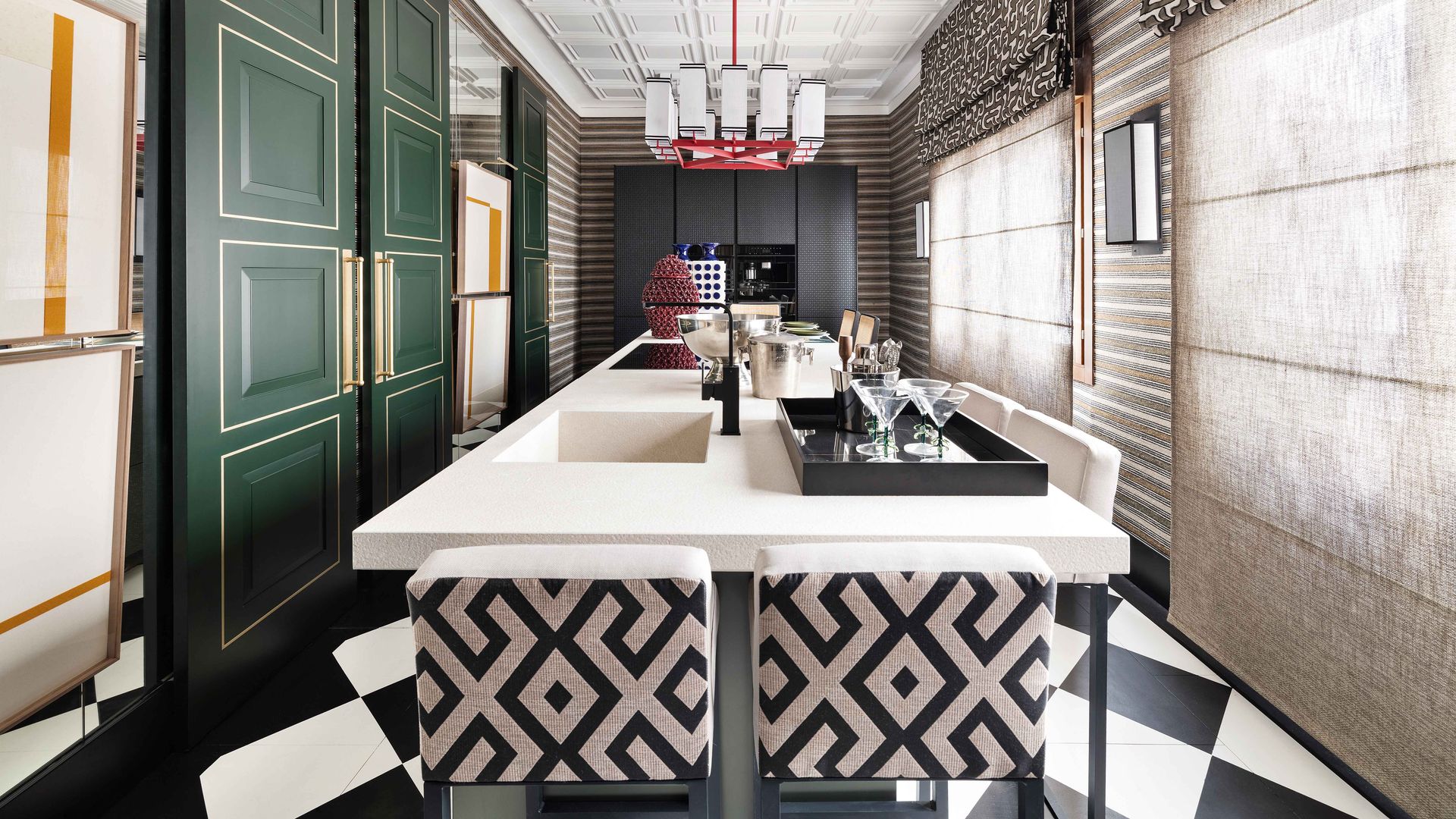 Cocina con isla central, suelo de damero blanco y negro, sillas con tela con motivos geométricos blancos y negros, estores, puertas verdes, molduras en el techo