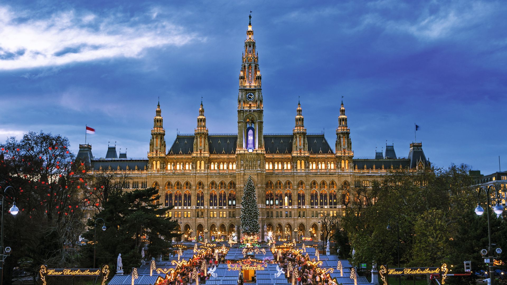 Plaza con multitud en mercado navideño frente a edificio iluminado al atardecer.
