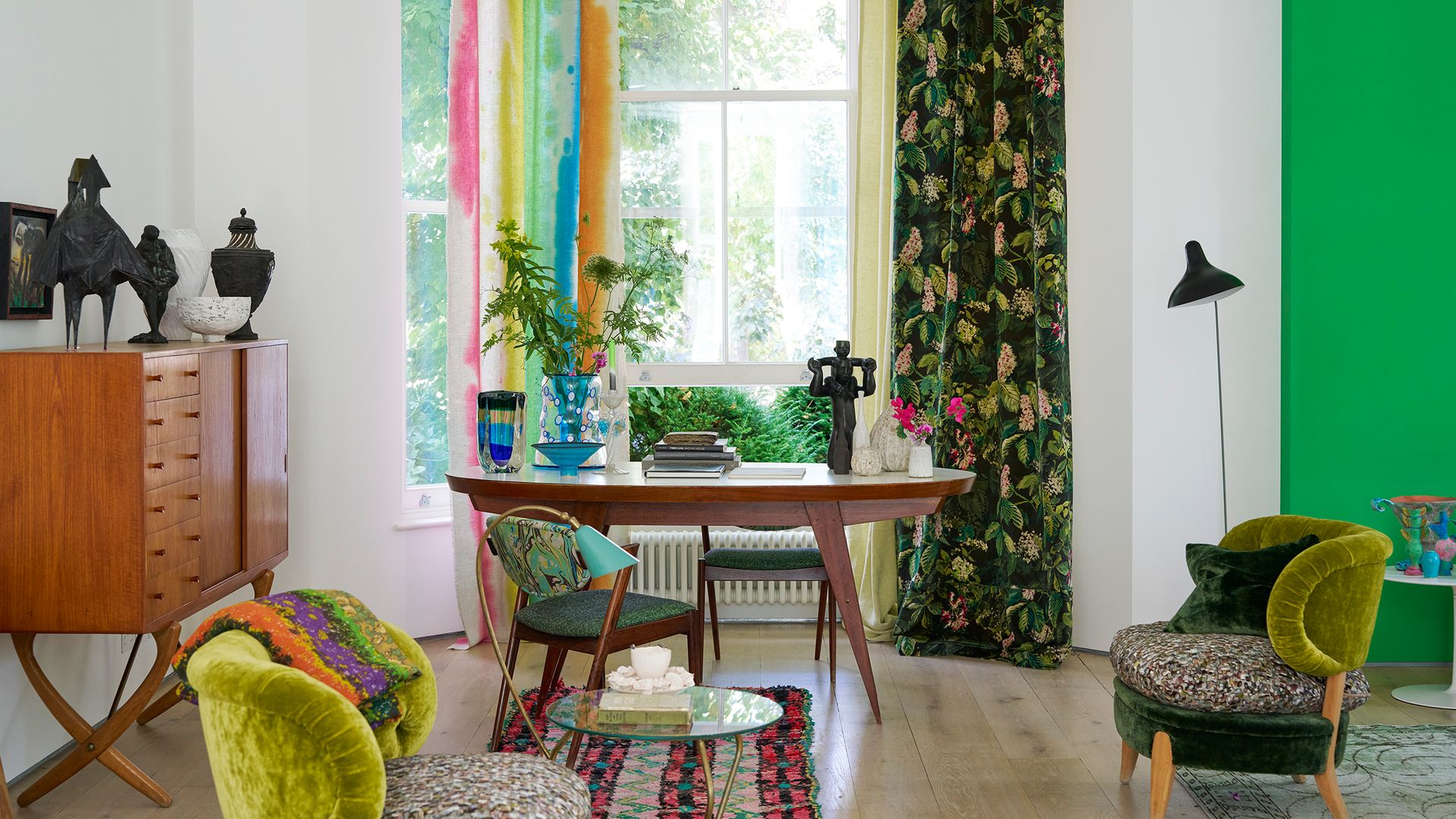 Despacho con mesa de madera oscura, cortinas de flores y de rayas, pared verde, aparador de madera, alfombra y butacas tapizadas