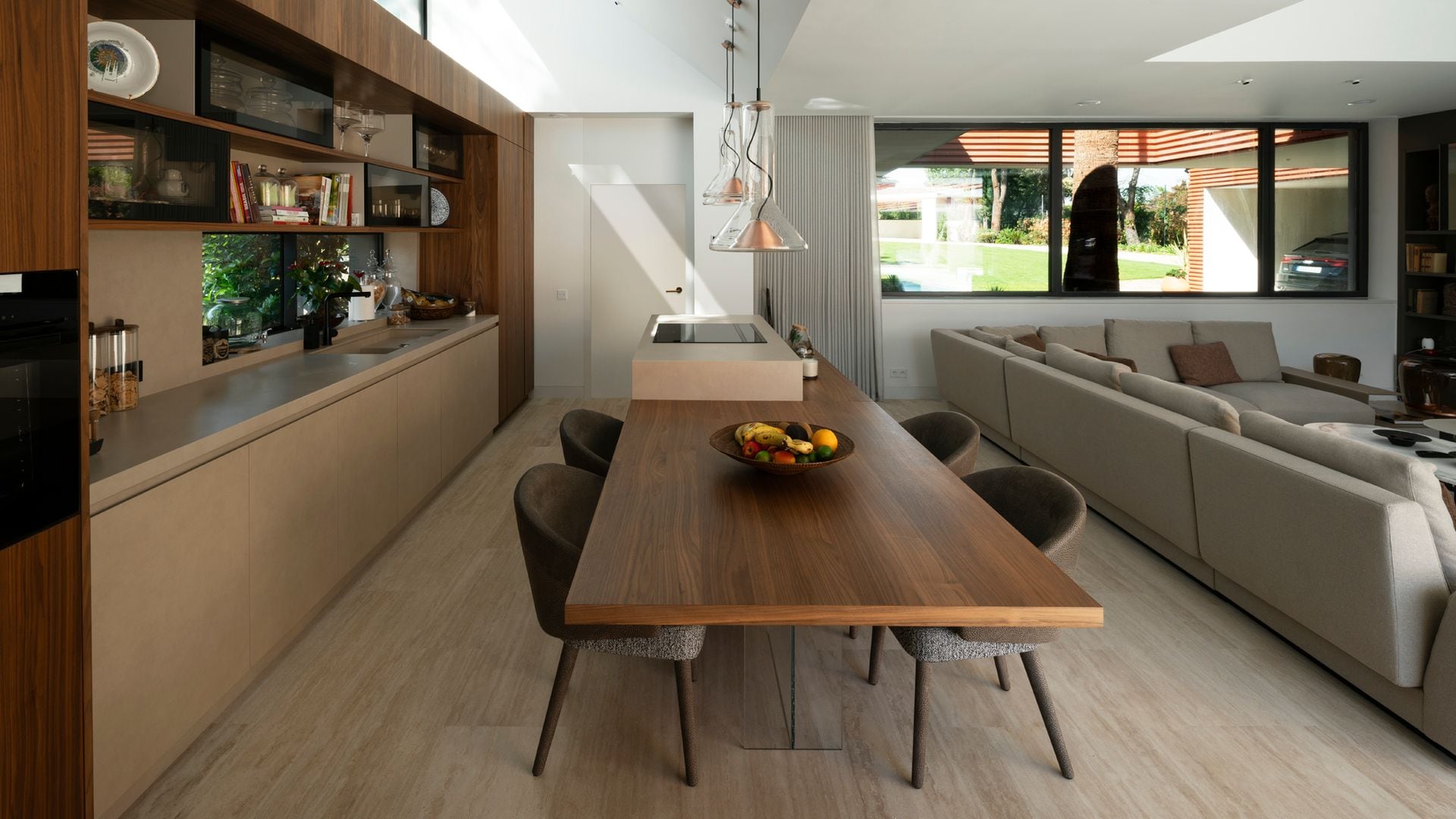 Cocina abierta con comedor, muebles color camel, gran mesa de madera adosada a la isla, sofá XL de color gris, lámparas de techo 