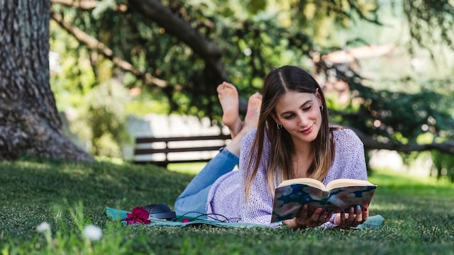 Chica leyendo tumbada en el césped de un parque, al lado de un árbol, con ropa de primavera y descalza
