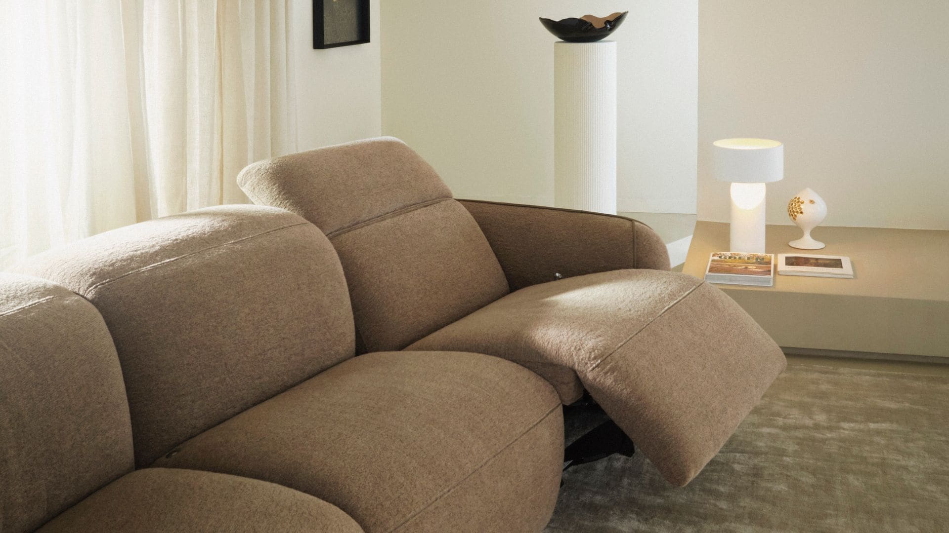 Sofá en color beige con asiento reclinable, cortinas blancas y lámpara de sobremesa