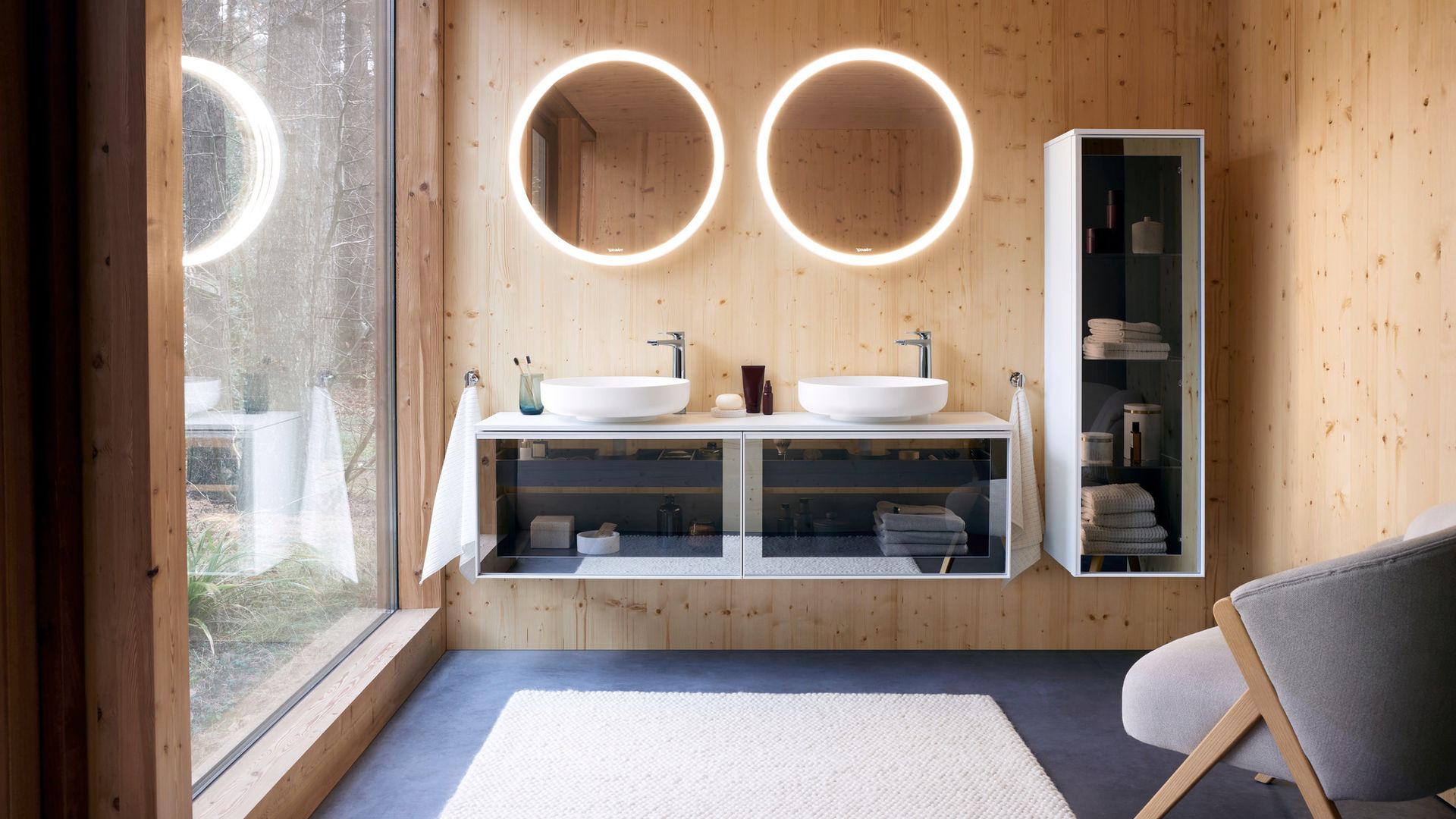 Baño con paredes de madera, dos lavamanos redondos sobre la encimera, mueble de lavabo con la puerta de cristal transparente, espejos redondos retroiluminados, columna con puerta de cristal suspendida