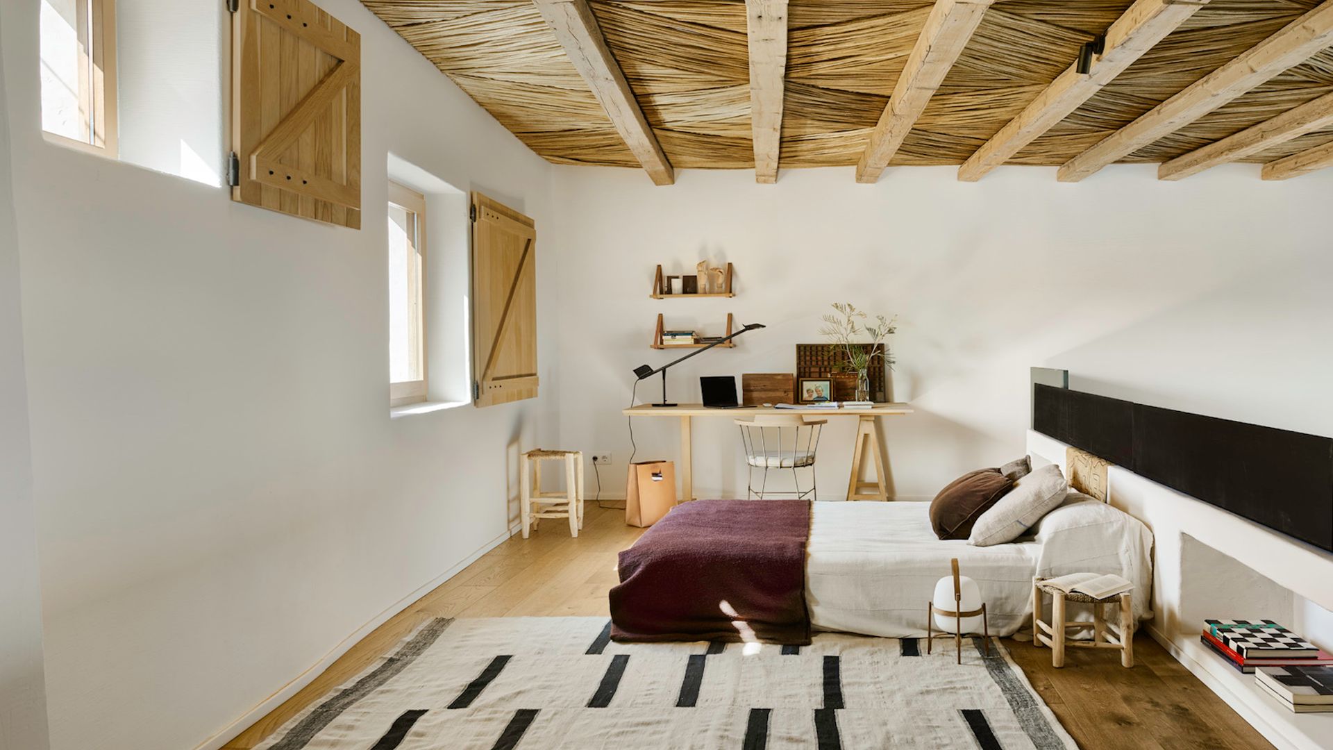 Dormitorio con vigas de madera, cabecero de obra con baldas, escritorio con mesa de madera, baldas, silla de oficina, alfombra