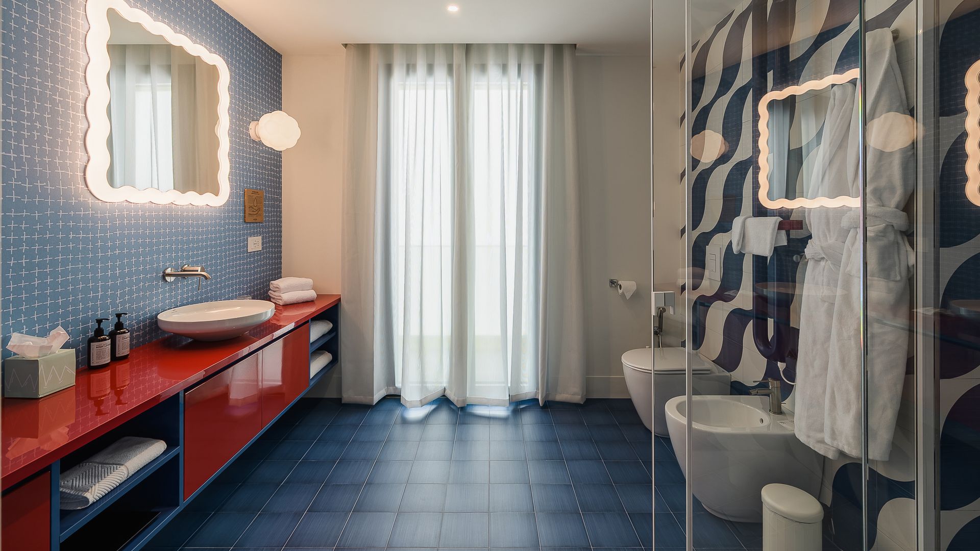 Baño grande en papel pintado con mueble bajolavabo rojo, suelo azul, espejo con luz, cortinas 