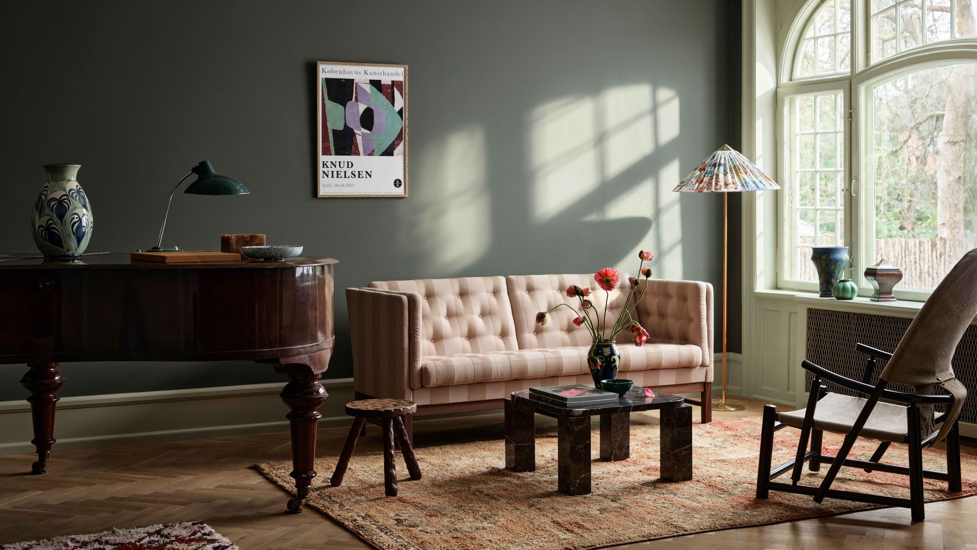 Salón con paredes gris oscuras, sofá con capitoné beige, gran ventanal, alfombra, molduras en el techo, mueble de caoba oscura