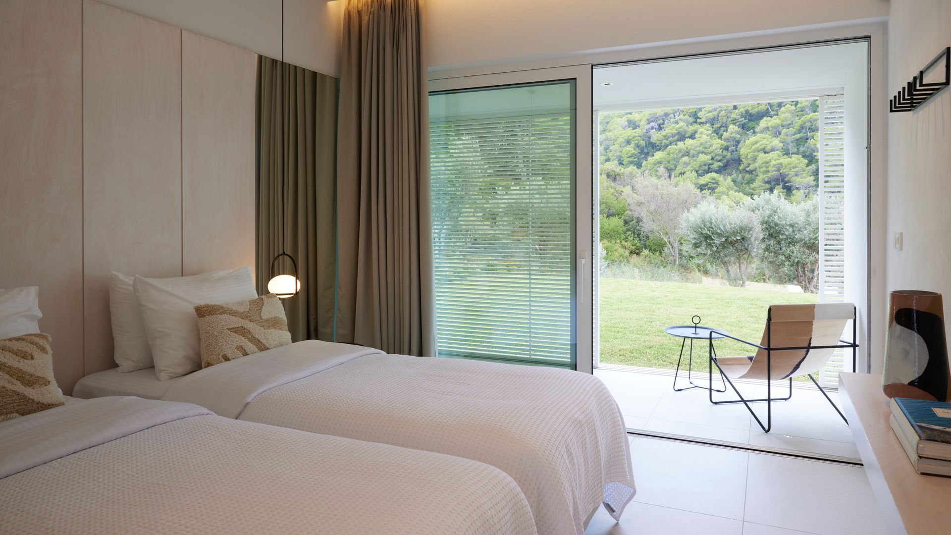 Dormitorio con dos camas, salida al jardín y terraza con hamaca y mesa, pared del cabecero de madera, cortinas beiges oscuro y lámpara de techo a ambos lados de la cama 