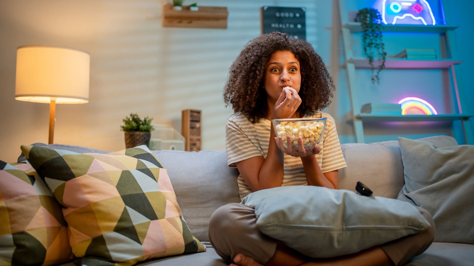 Chica joven sentada en el sofá de su salón con un bol de palomitas entre las manos y mirando hacia la televisión.