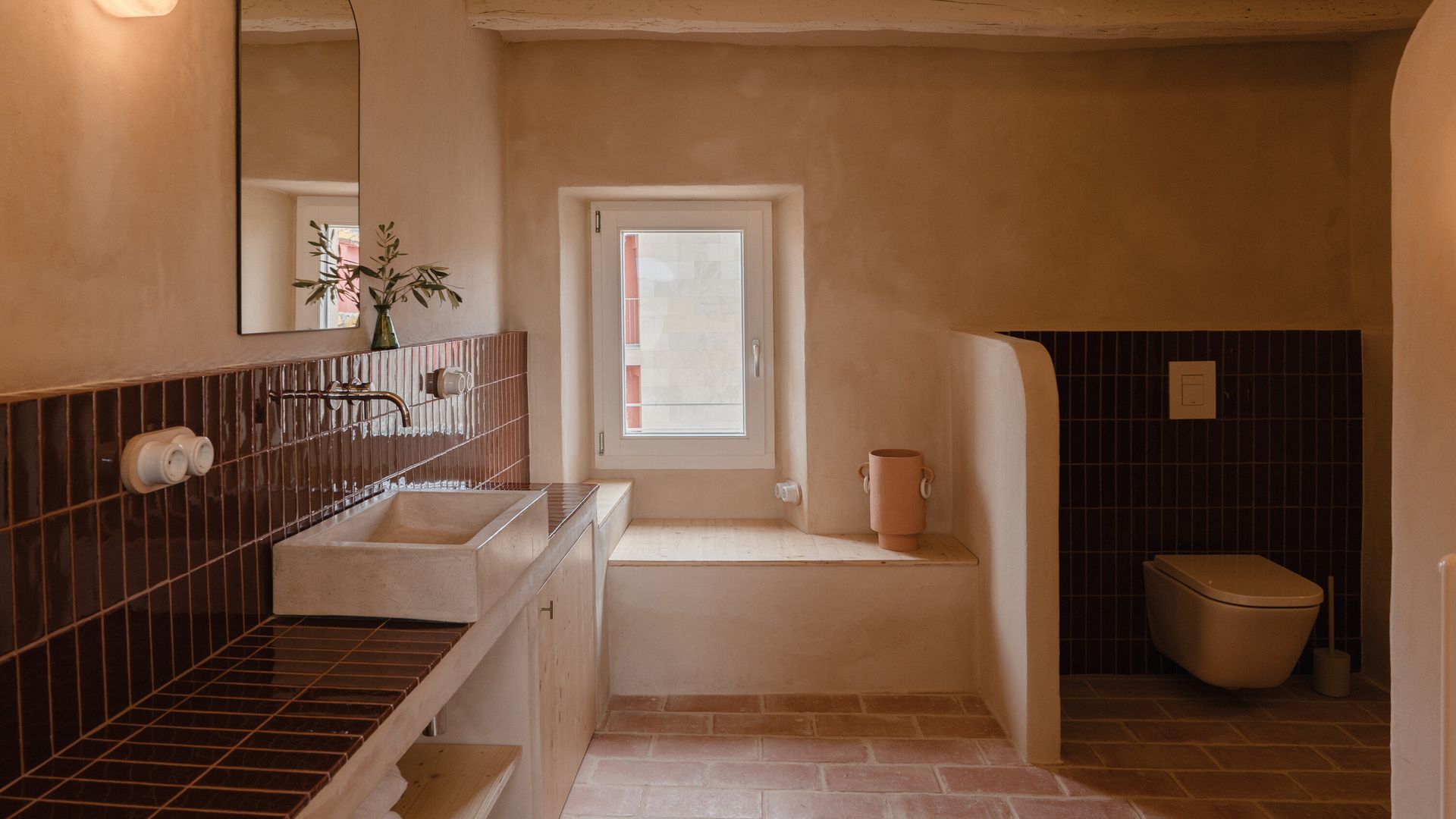 Baño con murete de obra separando el sanitario, encimera y antepecho de azulejos marrones brillantes, espejo, aplique de pared