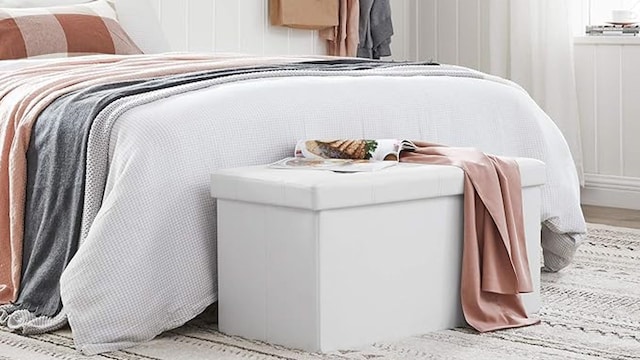 Puf de almacenaje SONGMICS de Amazon en un dormitorio decorado en blanco, colocado a los pies de la cama.