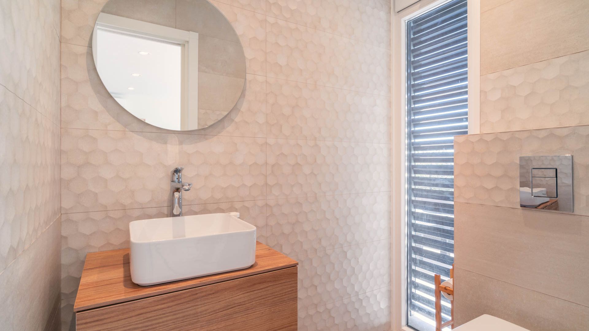 Baño con revestimiento de azulejos beige, espejo redondo, mueble bajolavabo de madera, ventana con salida a terraza