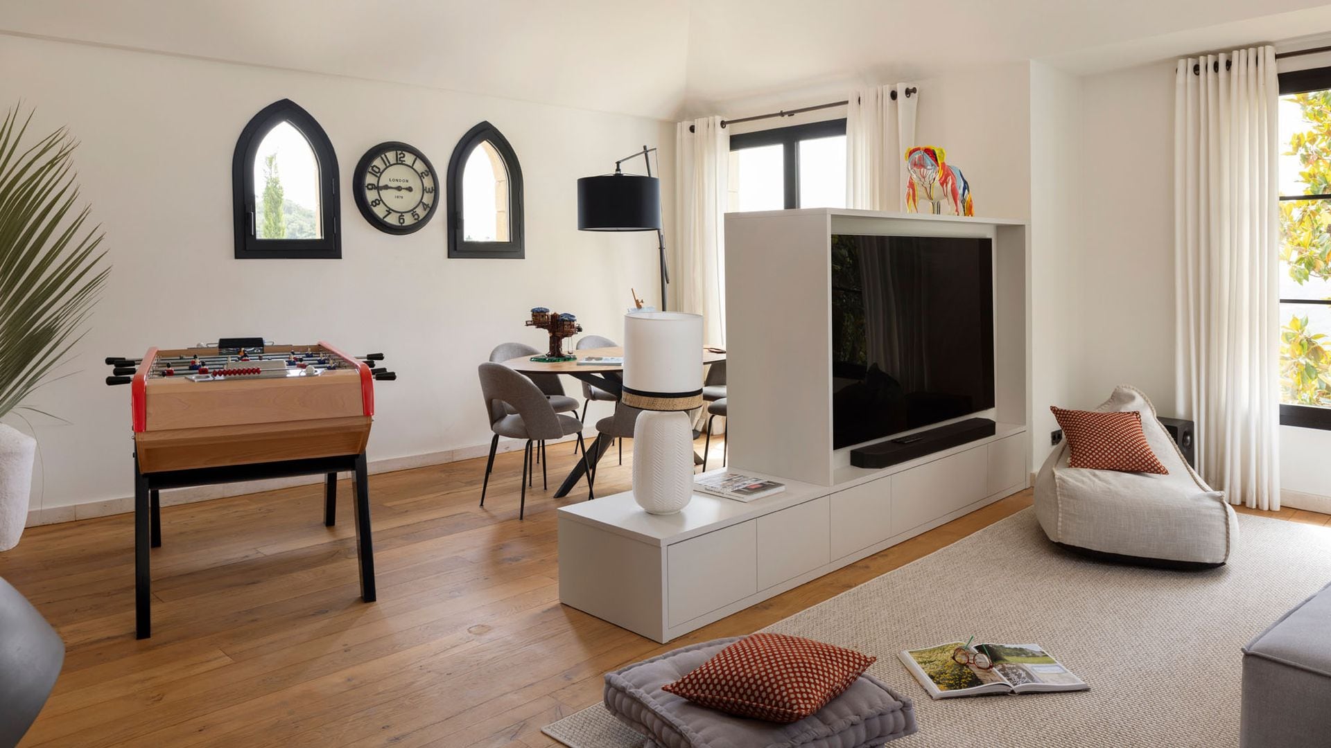 Salón con zona de juegos con futbolín y mesa redonda, con mueble con televisión blanco que separa el estar de la zona de juegos, pufs, cojines de suelo, alfombra