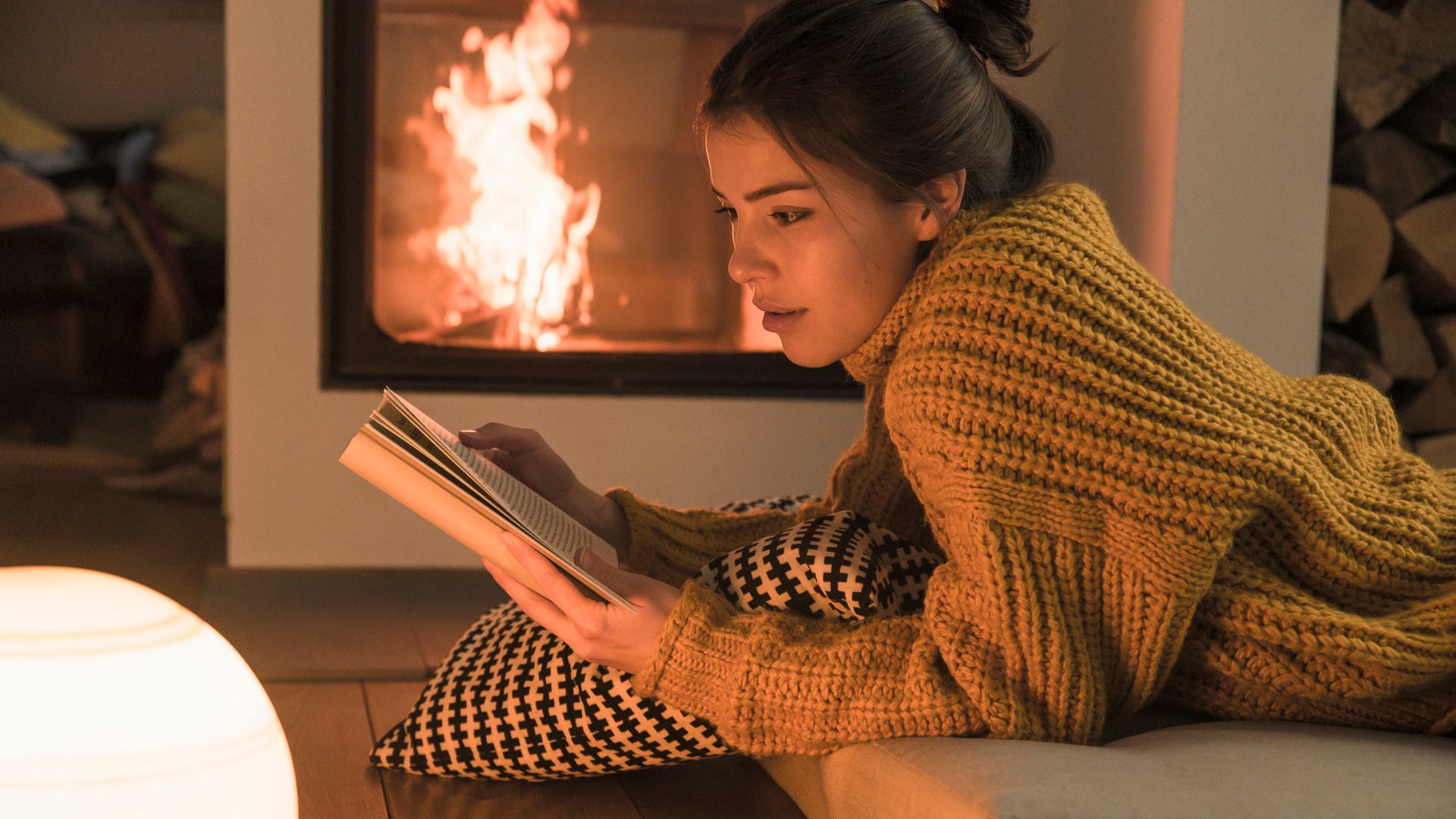 Mujer leyendo un libro sobre un cojín al lado de la chimenea con un jersey gordo en color mostaza.