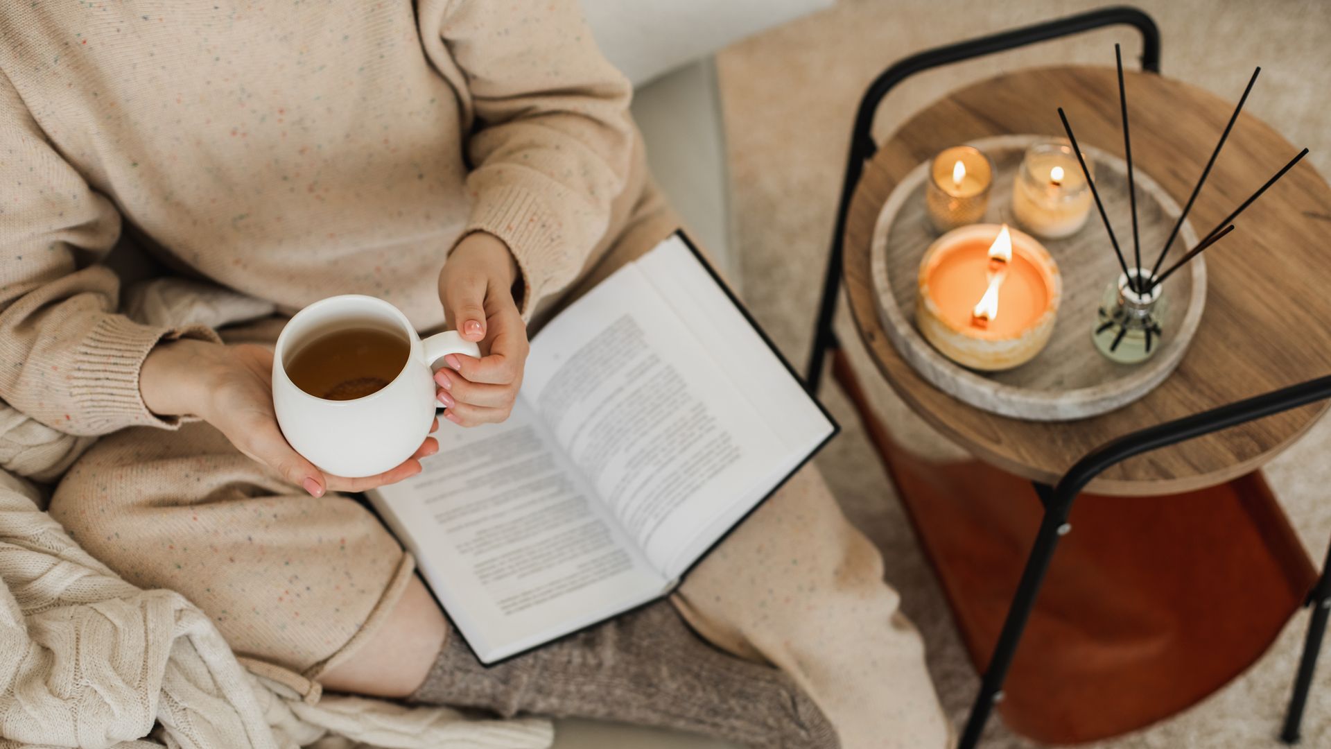 Chica leyendo en casa, sentada en el sofá, con ropa de otoño-invierno, velas encendidas y una infusión entre las manos, en una taza blanca. Plano desde arriba.