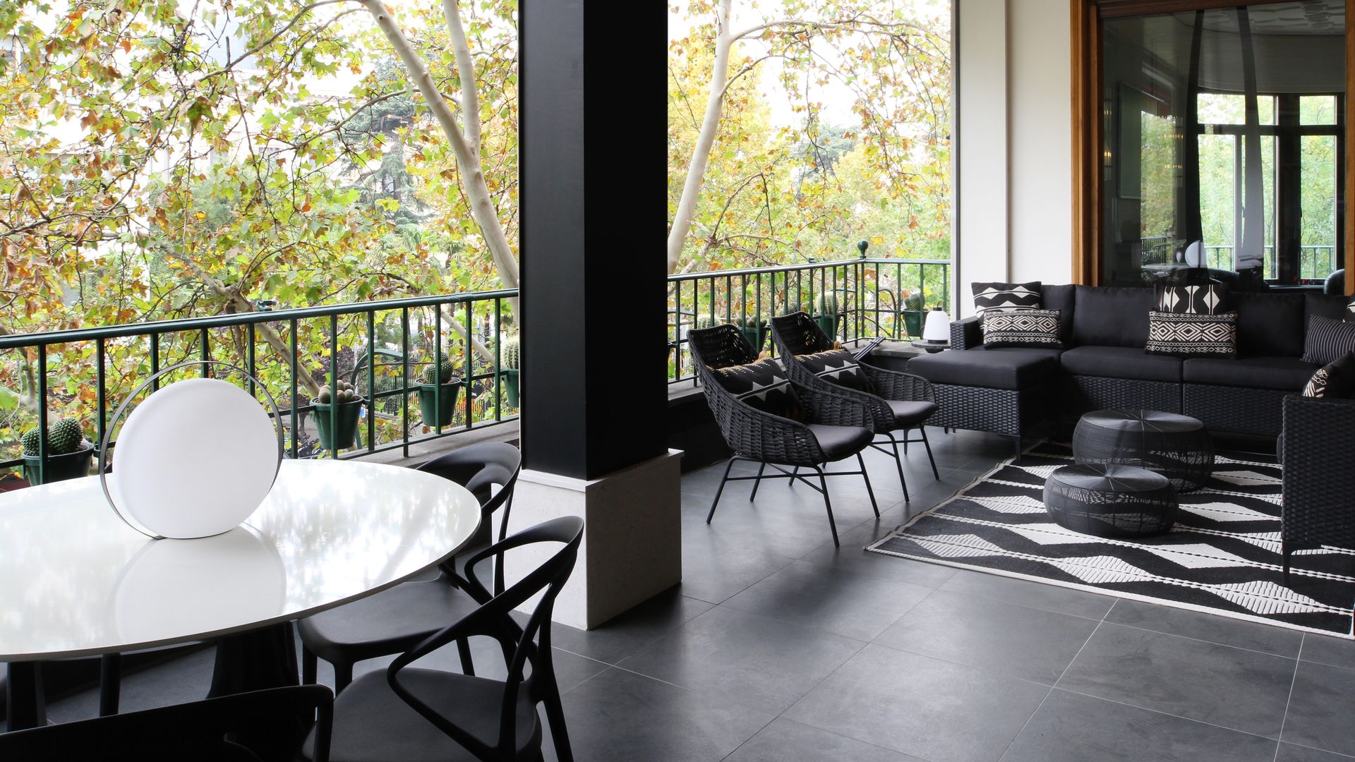 Terraza con suelo de baldosas negras, zona de estar con sofá negro, alfombra geométrica y butacas negras y una zona de comedor con mesa redonda blanca y sillas negras 