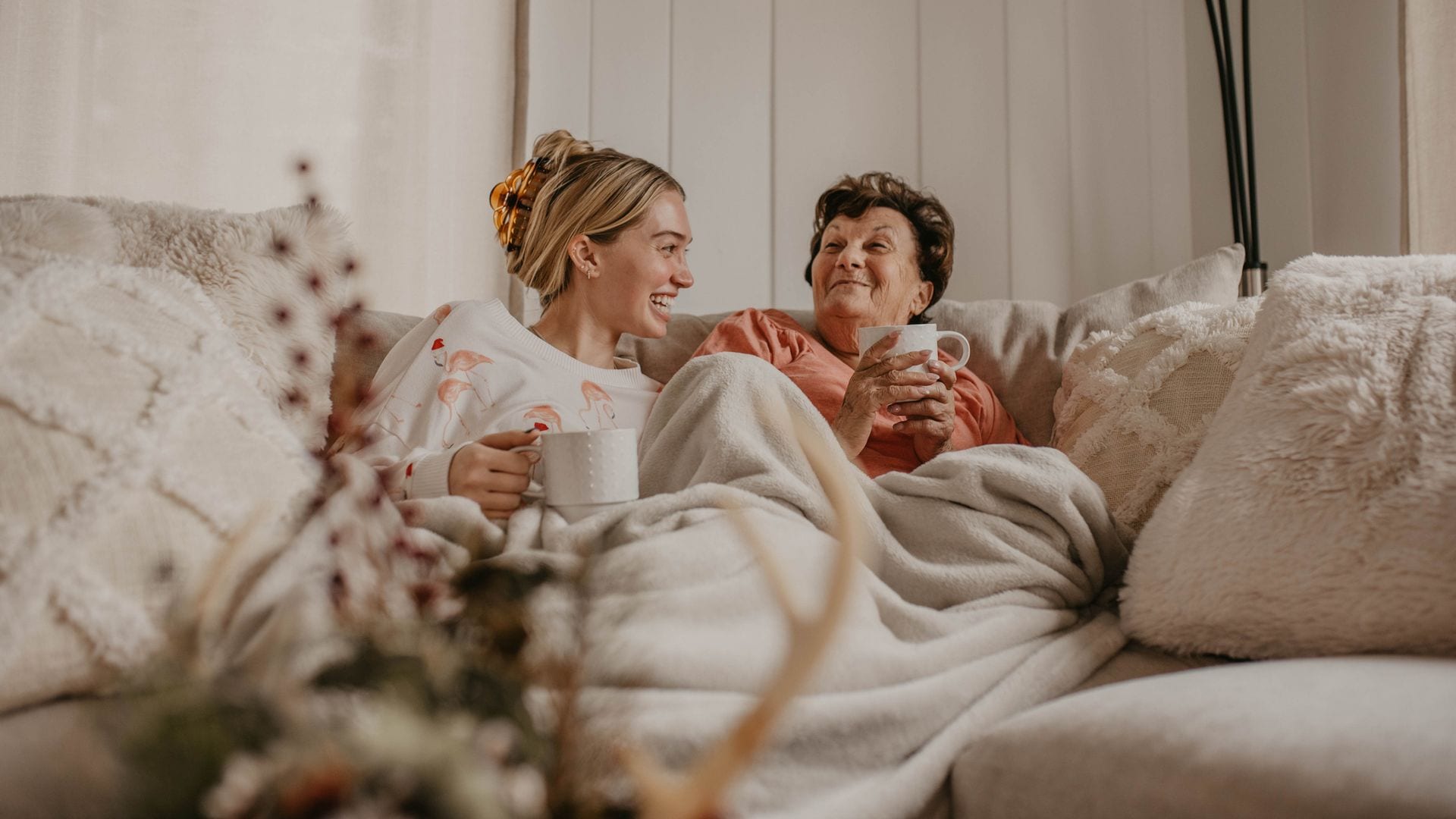 Abuela y nieta en el sofá de casa con una manta en invierno, disfrutando de una taza de café mientras ríen.