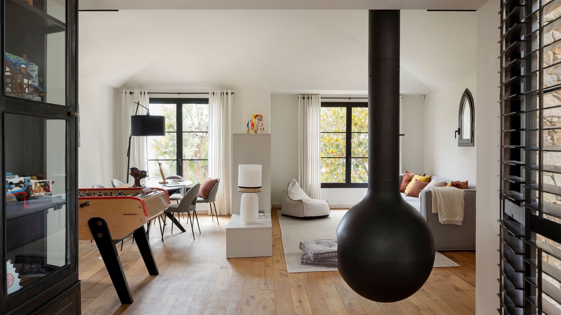 Salón comedor con chimenea colgada del techo, suelo de madera, futbolín, sofá gris, mesa de comedor, mueble blanco entre ambos espacios
