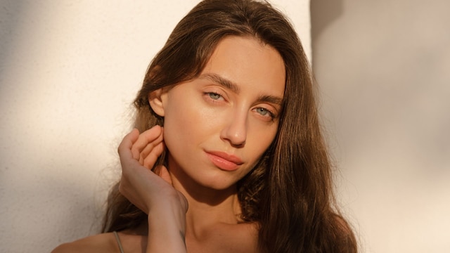 Mujer con cabello castaño largo y ojos azules apoyada en una pared con el sol en el rostro