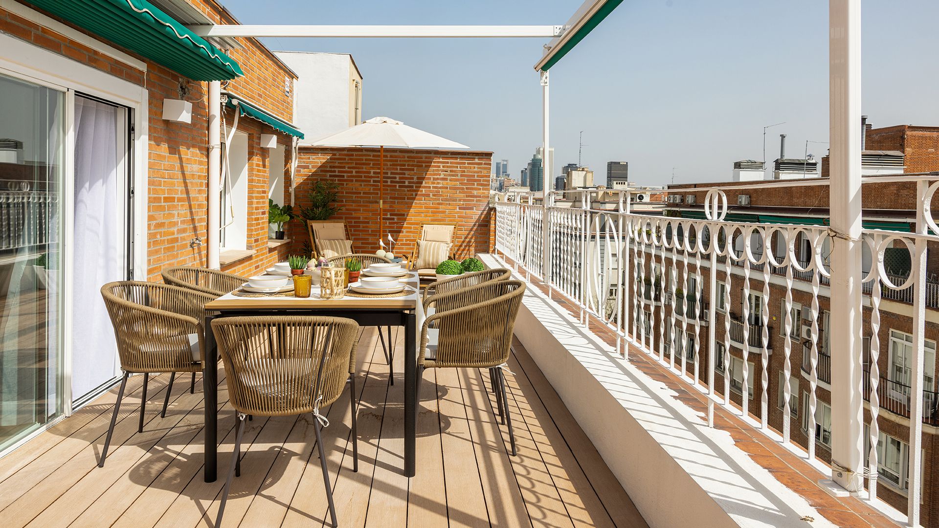 Terraza urbana con mesa de comedor y sillas de fibras vegetales, toldo verde, suelo de madera, barandilla blanca 