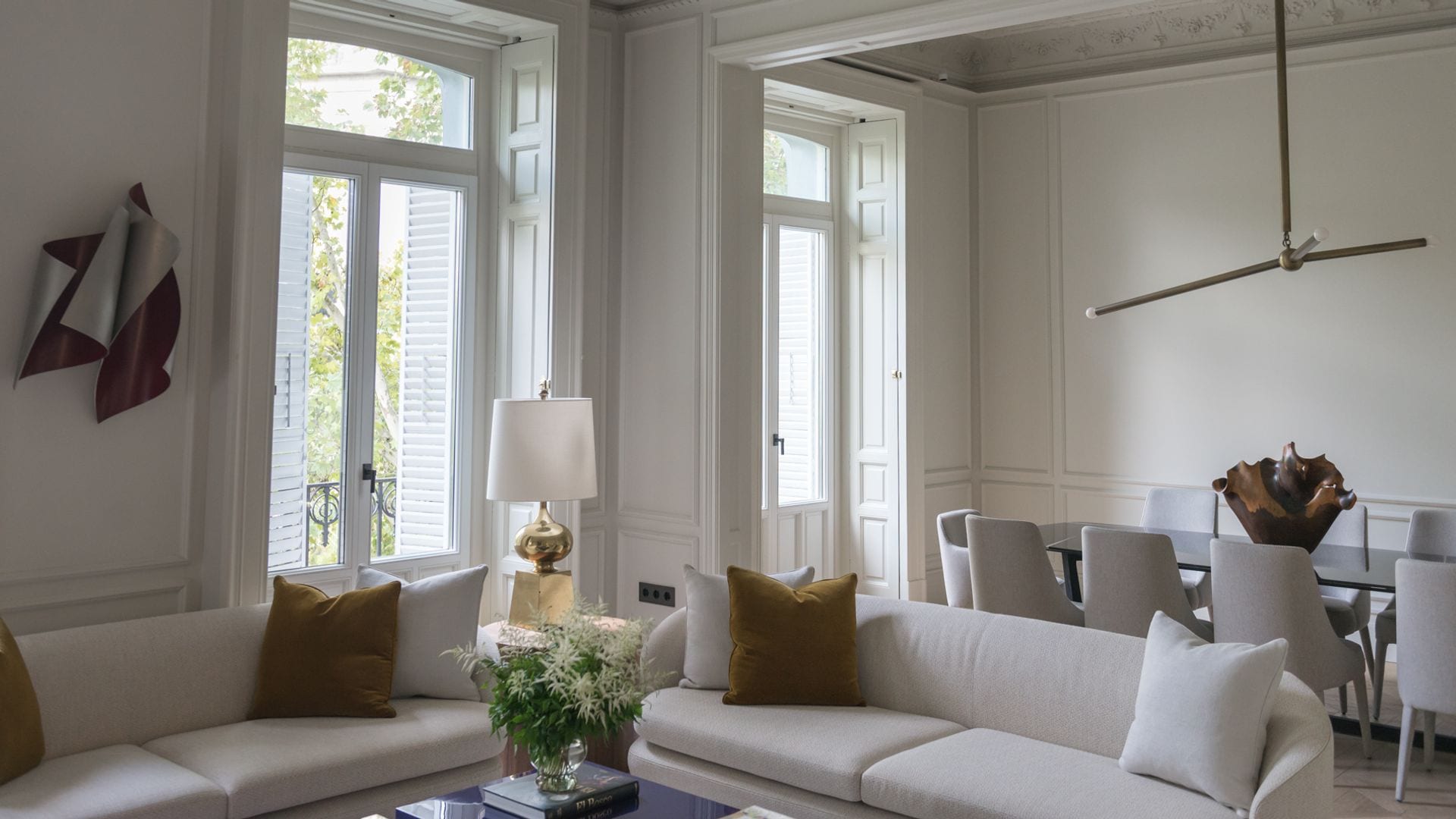 Salón comedor con dos grandes balcones, sofás blancos, mesa de comedor, sillas tapizadas, lámpara de techo en la mesa de comedor, lámpara de sobremesa al lado del sofá