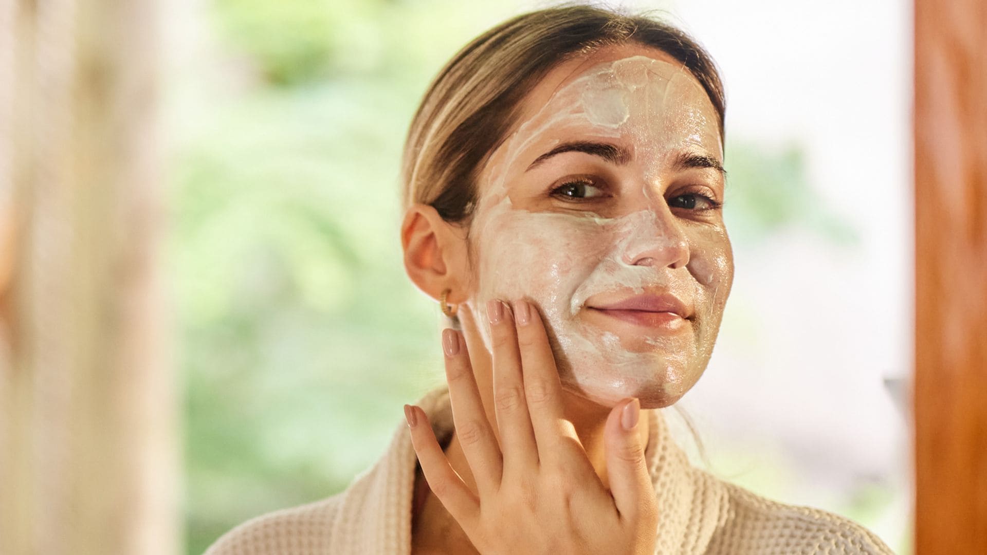 Mujer con bata de ducha mirándose al espejo con mascarilla de arcilla en el rostro