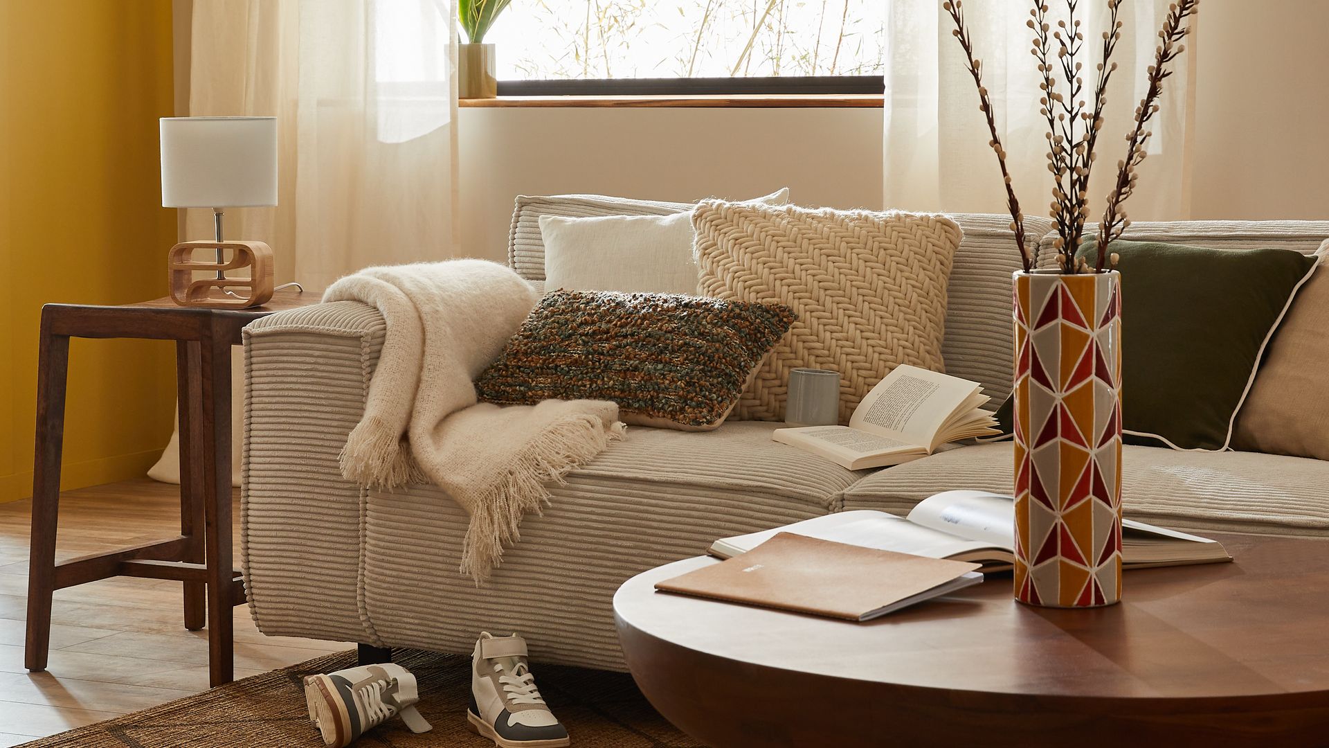 Salón con lámpara de techo de fibras vegetales, ventana, cortinas beiges, sofá beige, zapatillas deportivas en la alfombra de yute, manta, cojines, mesa de centro redonda, jarrón 