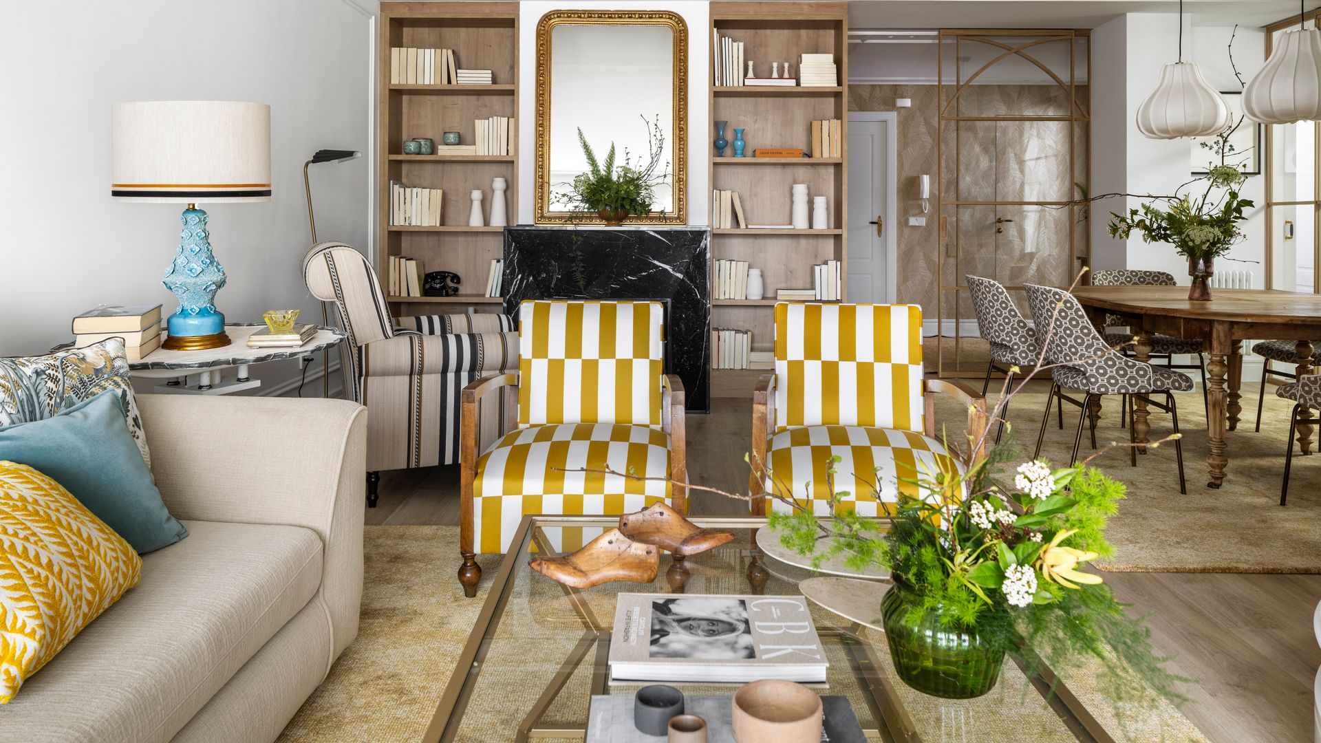 Salón comedor con chimenea, butacas moderas de rayas amarillas y blancas, sofá beige, alfombra, mesa de centro de cristal, mesa de comedor de madera con patas torneadas, lámparas de techo, sillas tapizadas, jarrón de cristal verde con flores, espejo dorado sobre la chimenea