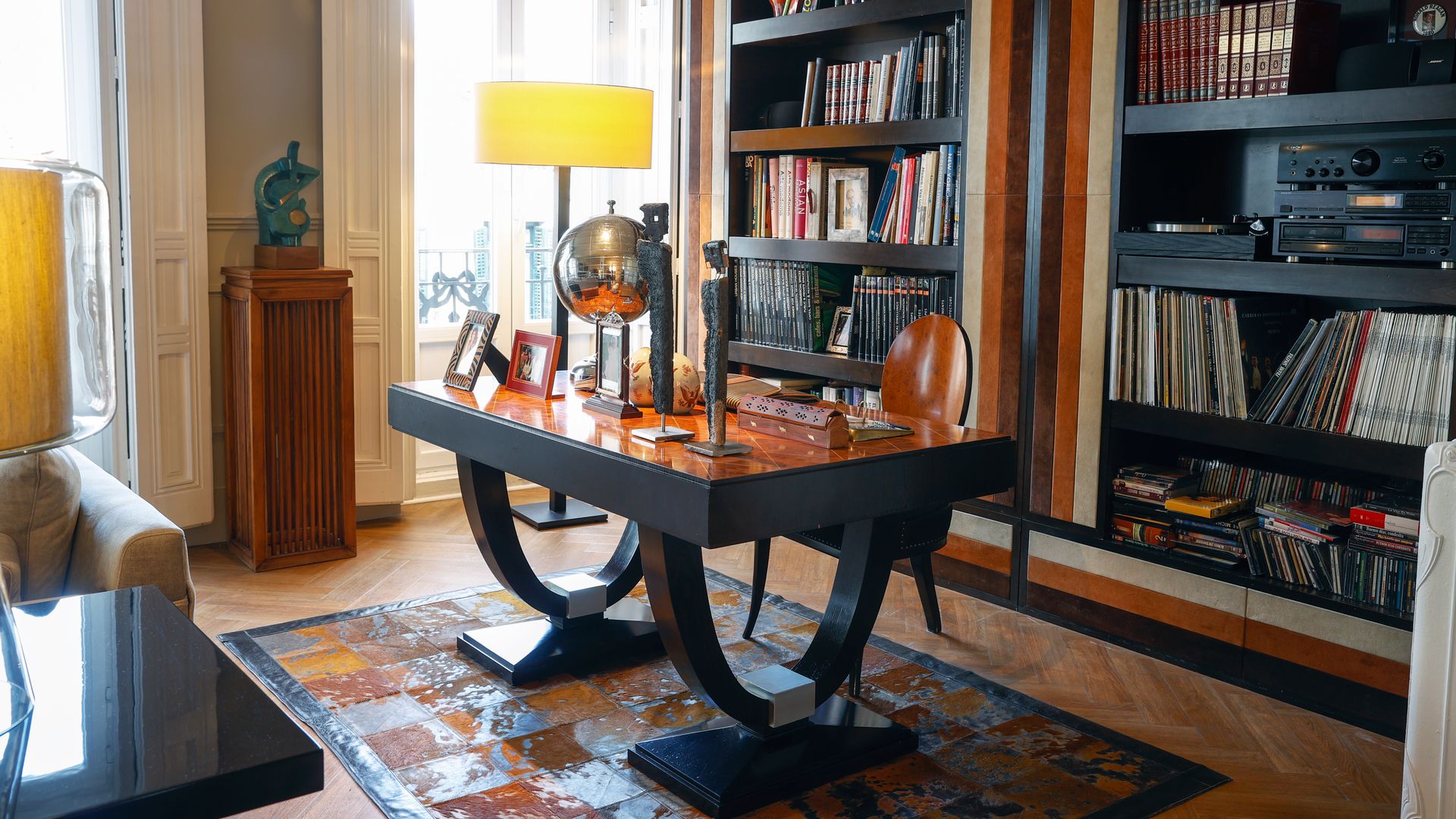 Despacho con mesa de madera, alfombra, librería de madera a medida, pedestal de madera con escultura, suelo de madera, silla de madera, lámpara de sobremesa