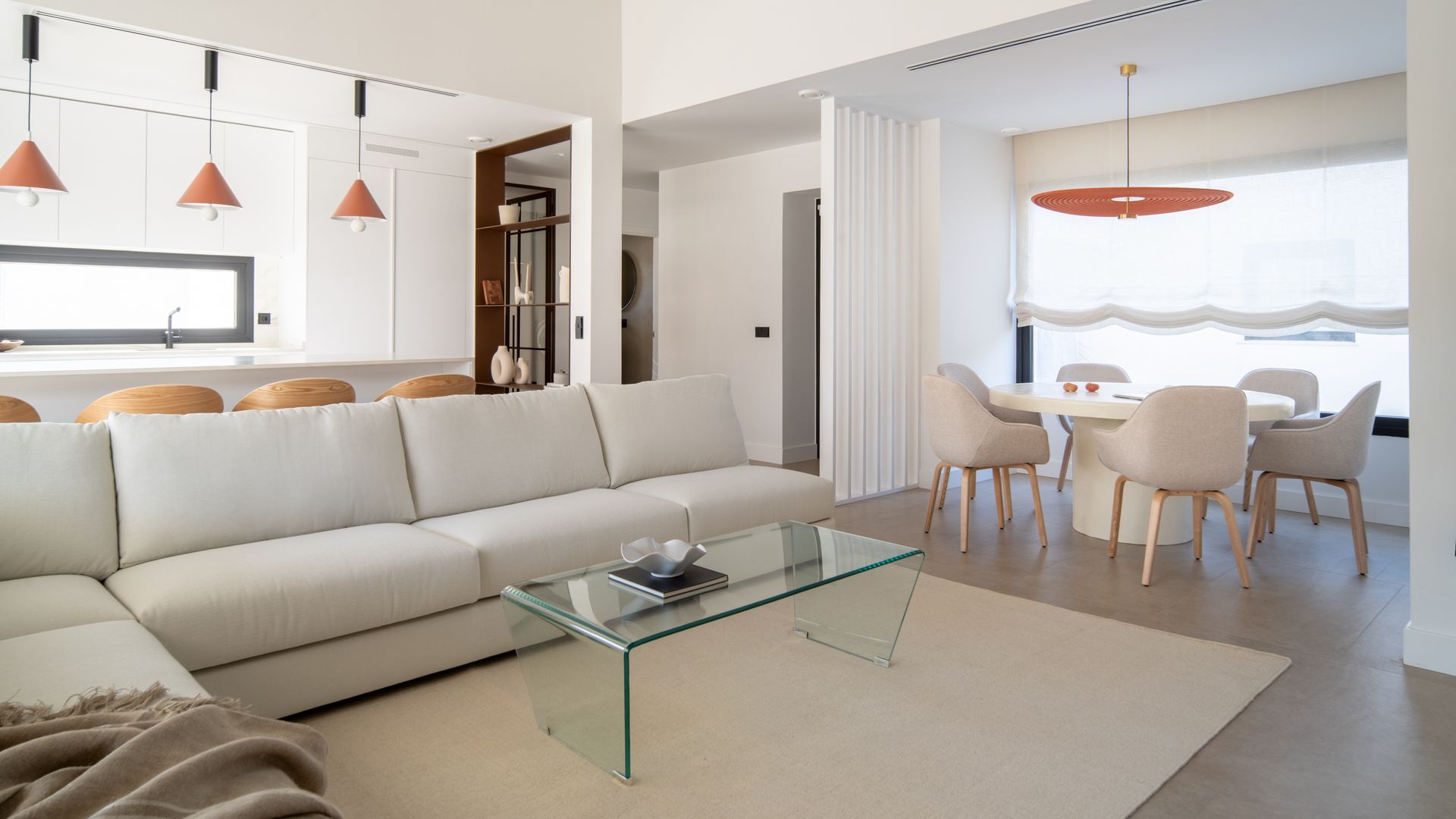 Salón comedor y cocina en color blanco, con detalles en madera y mesa de centro de cristal, lámparas de techo, alfombra, sofás blancos