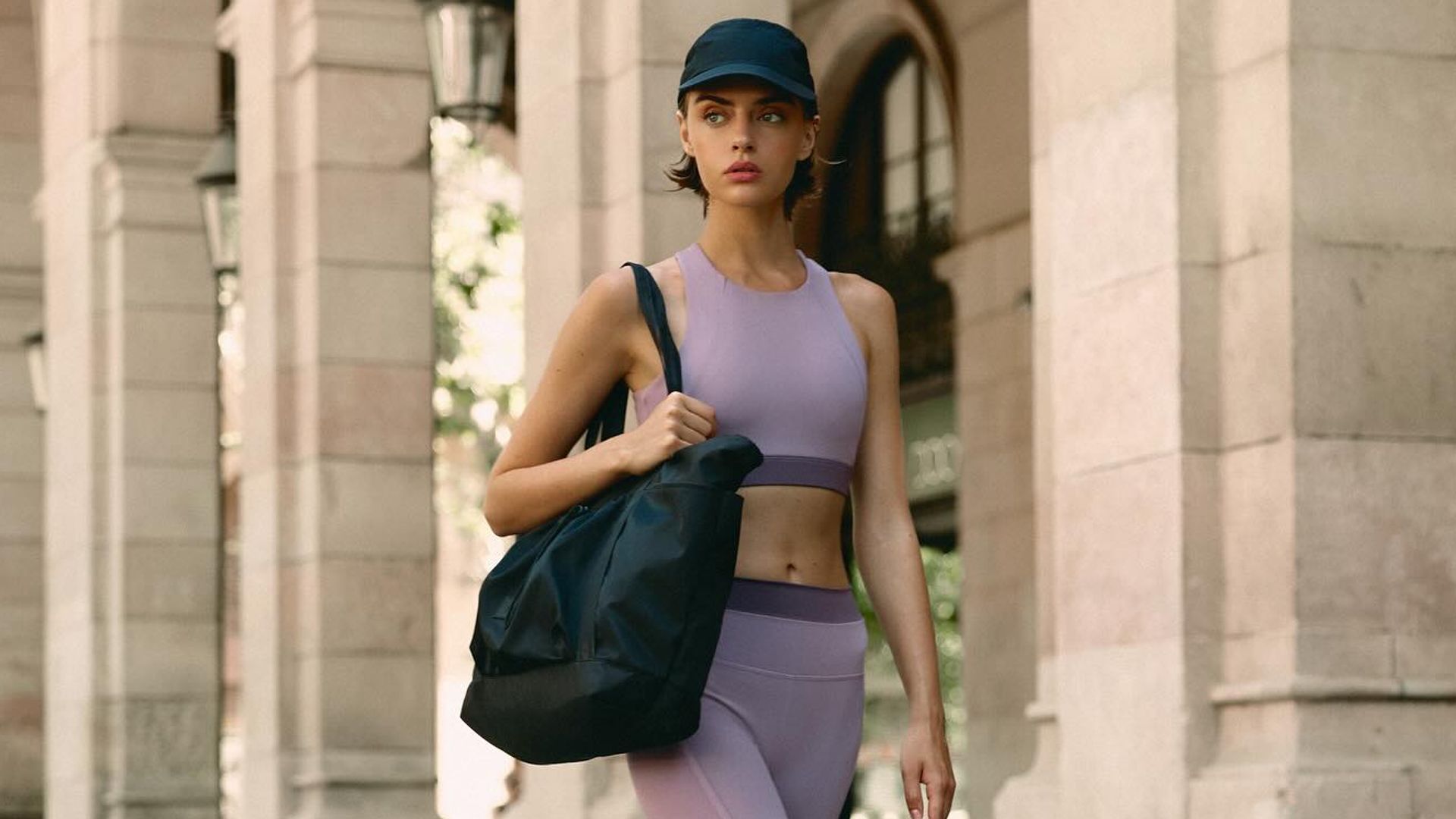 Mujer joven paseando por la calle con conjunto lila de Born, gorra y bolso negro de deporte